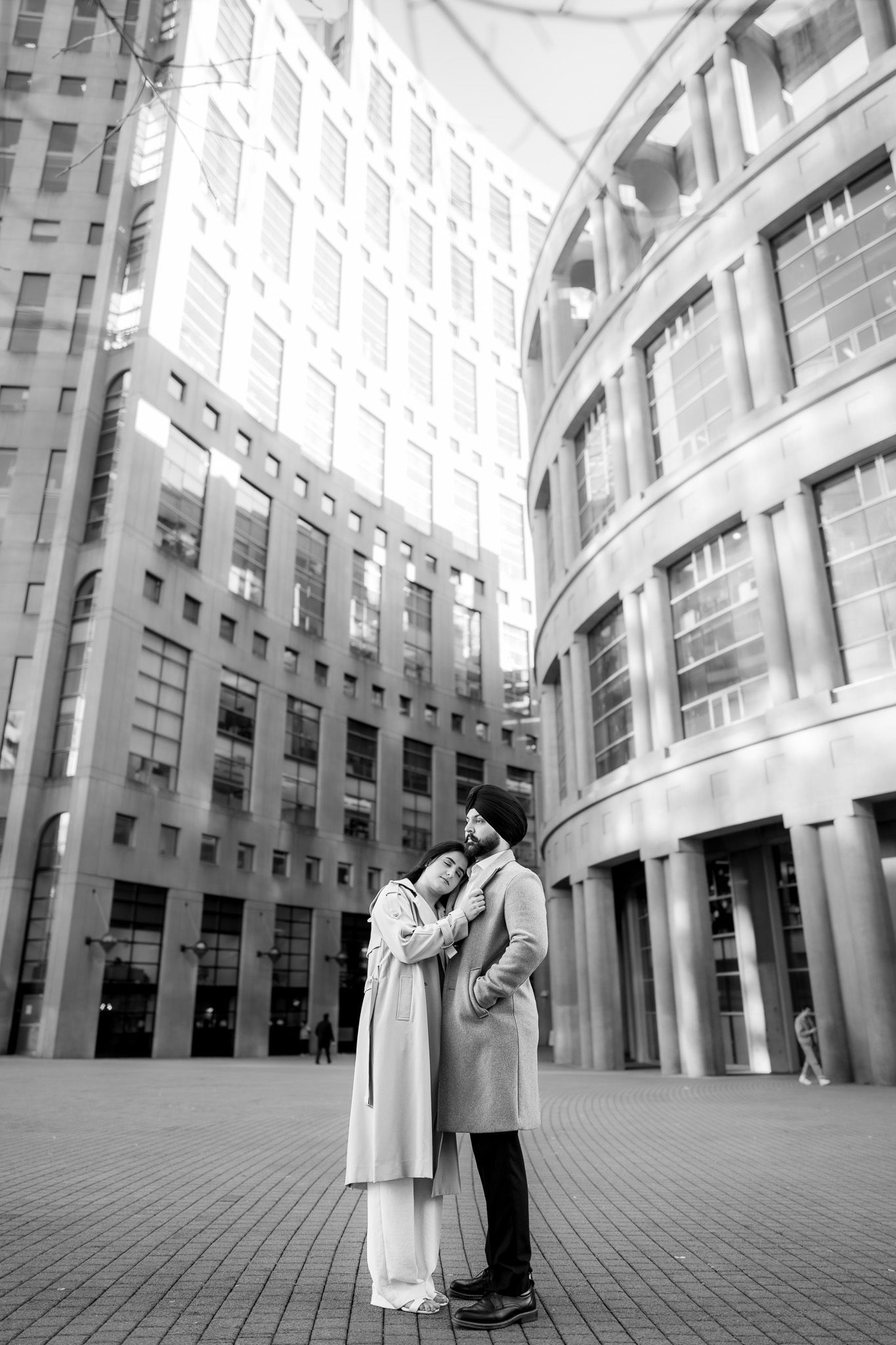A romantic black and white photo of a couple embracing in front of modern urban architecture with curved columns and buildings.