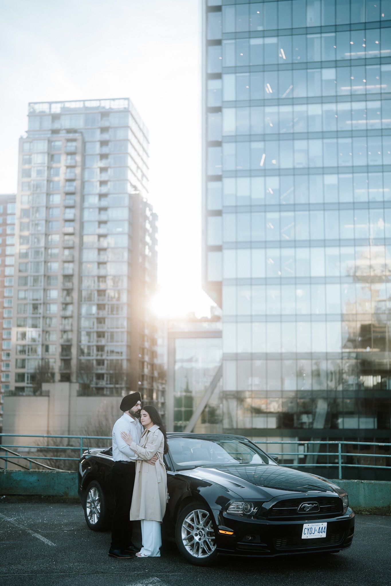 Romantic couple poses with sleek black BMW at sunset against modern glass building facade.