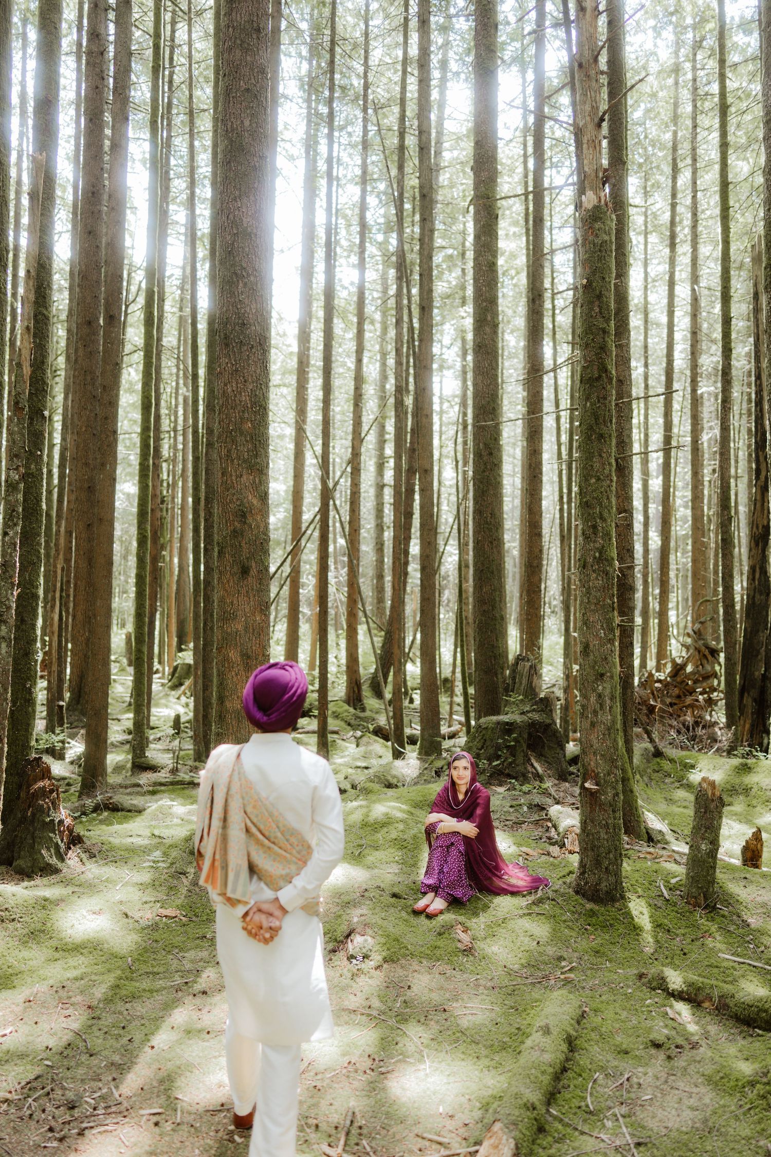Sikh couple in a sunlit forest, man in white kurta approaching woman seated on mossy ground among tall trees.