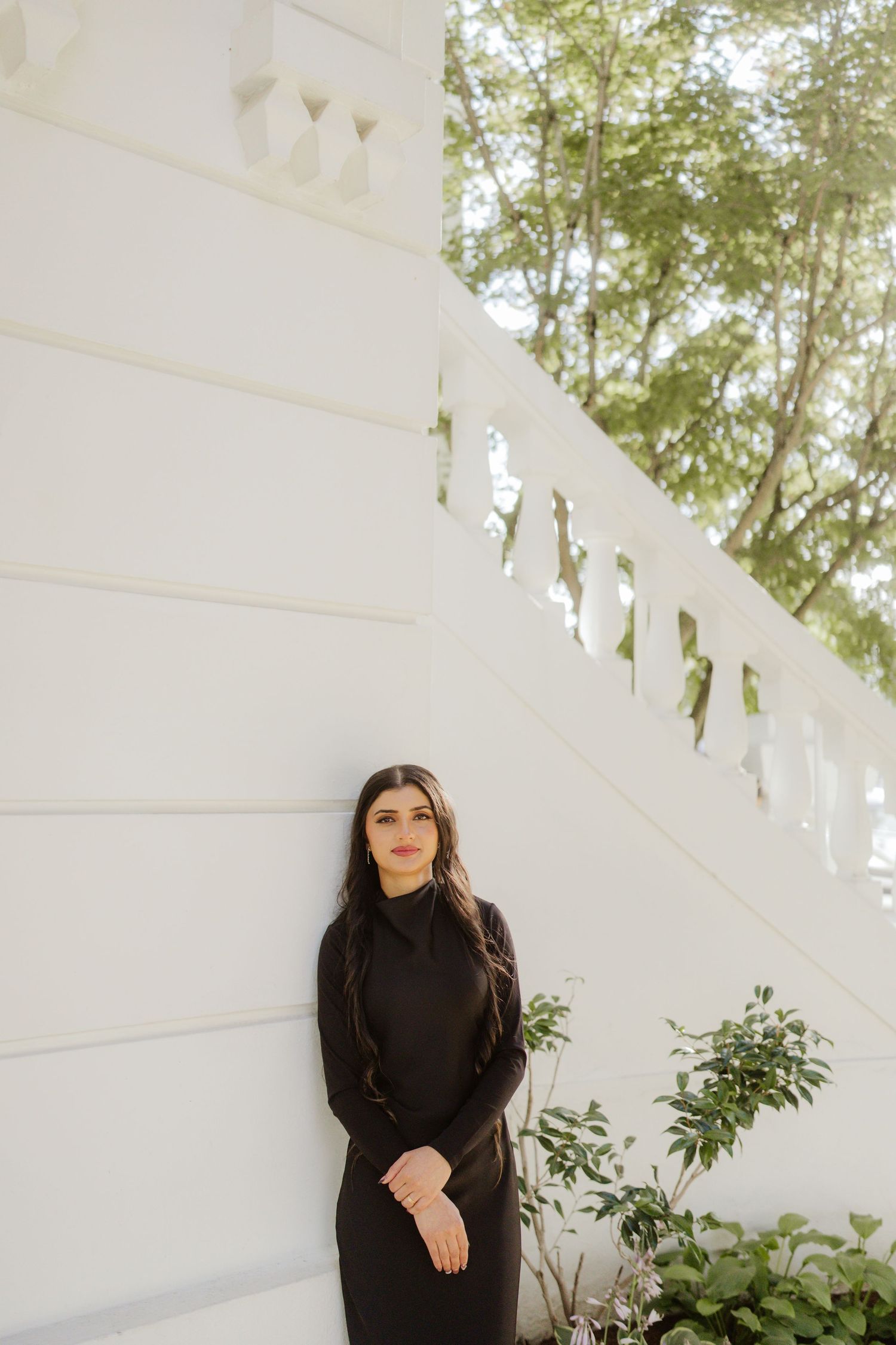 Elegant woman in black turtleneck dress leaning against a white decorative wall with green trees behind.