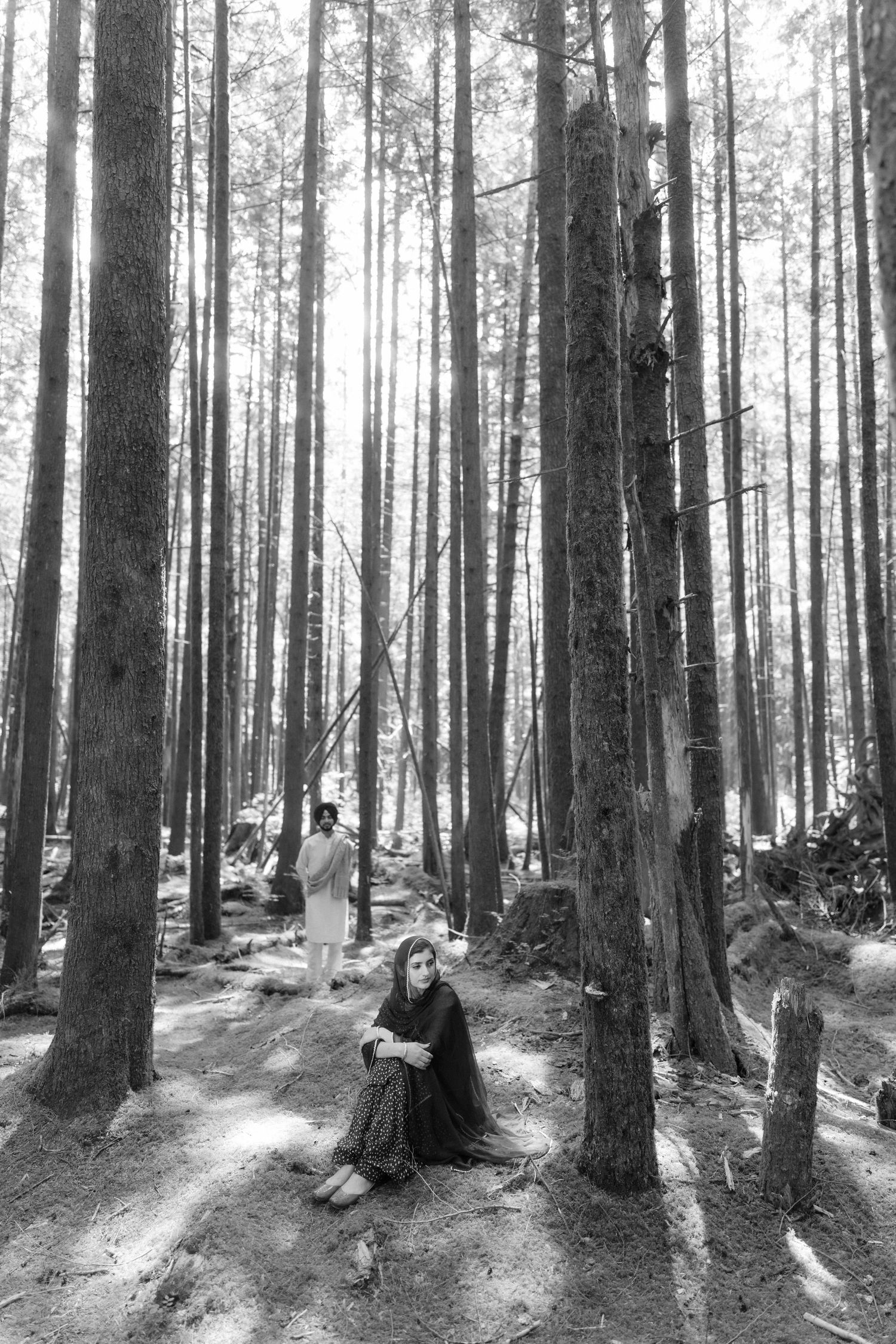 Black and white photo of a woman sitting on forest floor among tall pine trees with a person standing in the background.