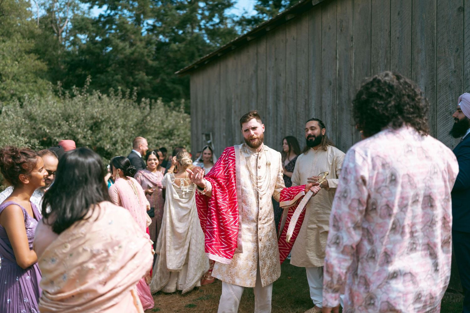 Groom in traditional Indian wedding attire with red dupatta walks with guests near a rustic barn.