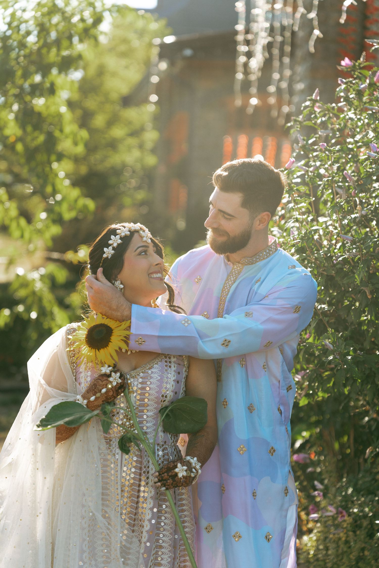 Couple in traditional South Asian wedding attire embrace outdoors, bride holding sunflower in white lehenga, groom in blue kurta.