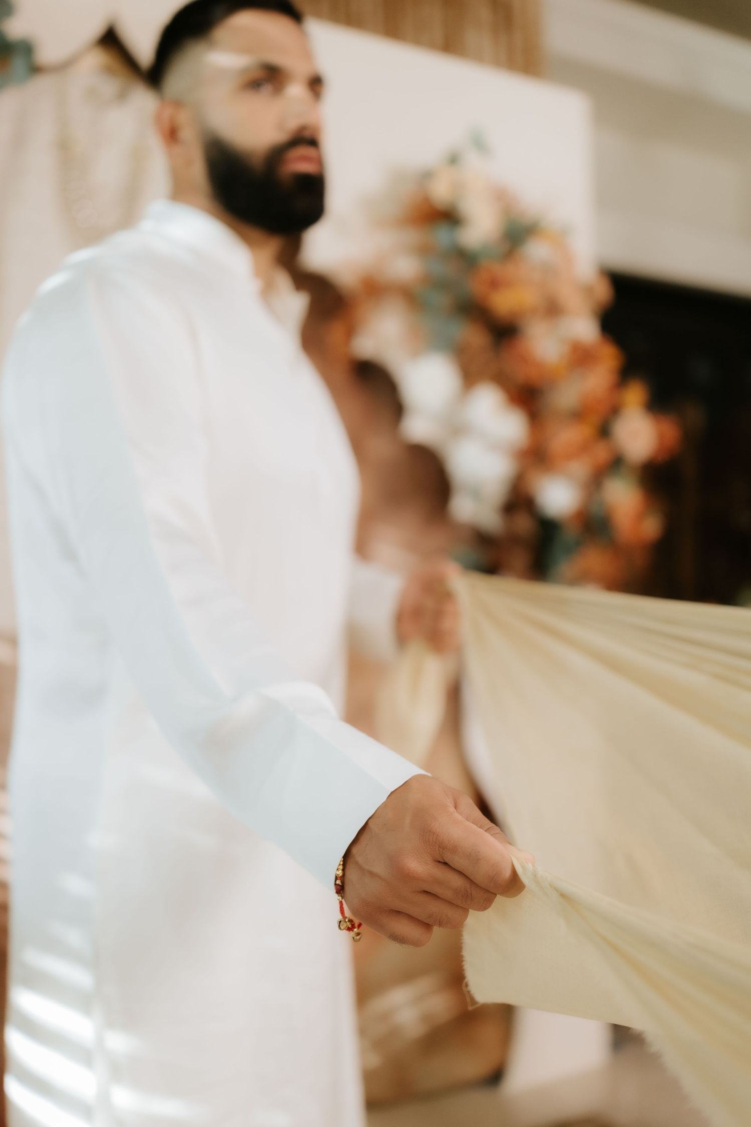Bearded man in white kurta holds flowing fabric at an elegant floral wedding ceremony.