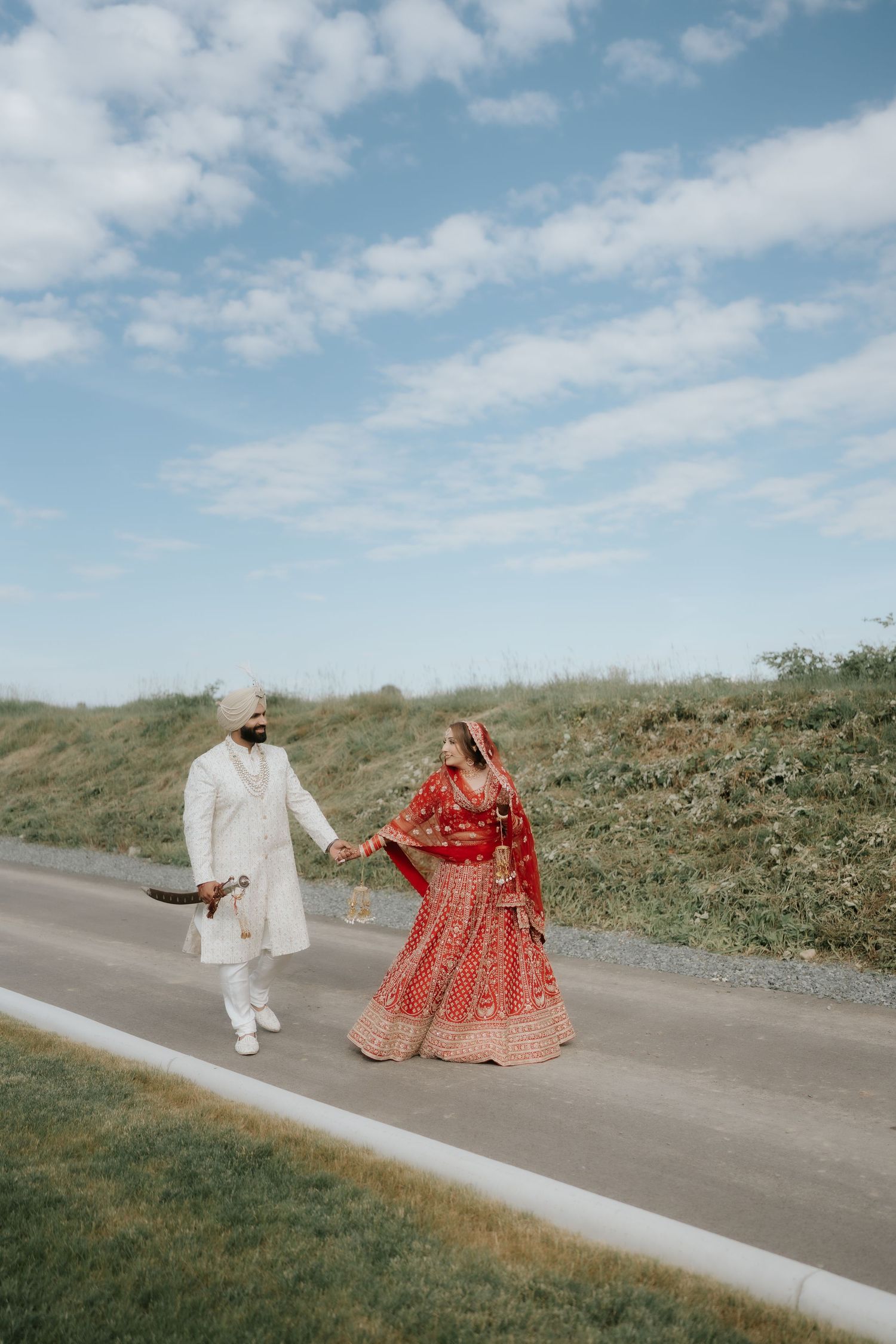 Indian bride in red lehenga and groom in white sherwani hold hands walking on a road under a blue sky.