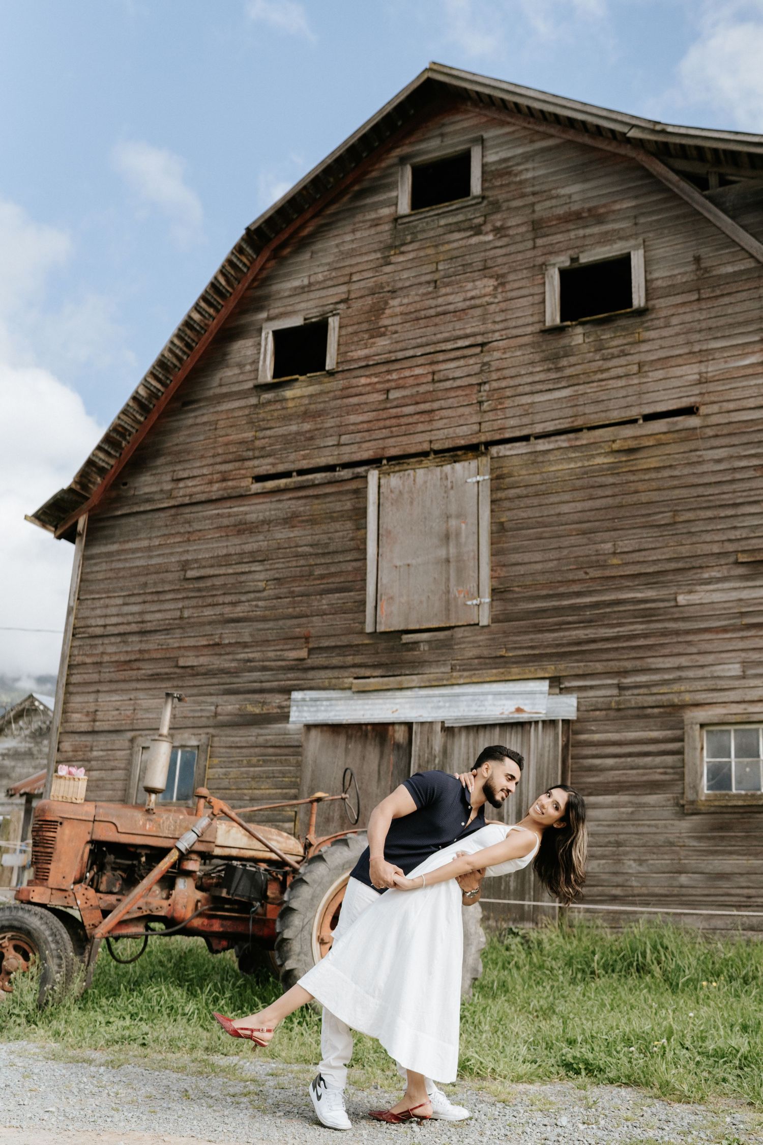 A couple poses romantically in front of a rustic wooden barn with an old red tractor in the background.