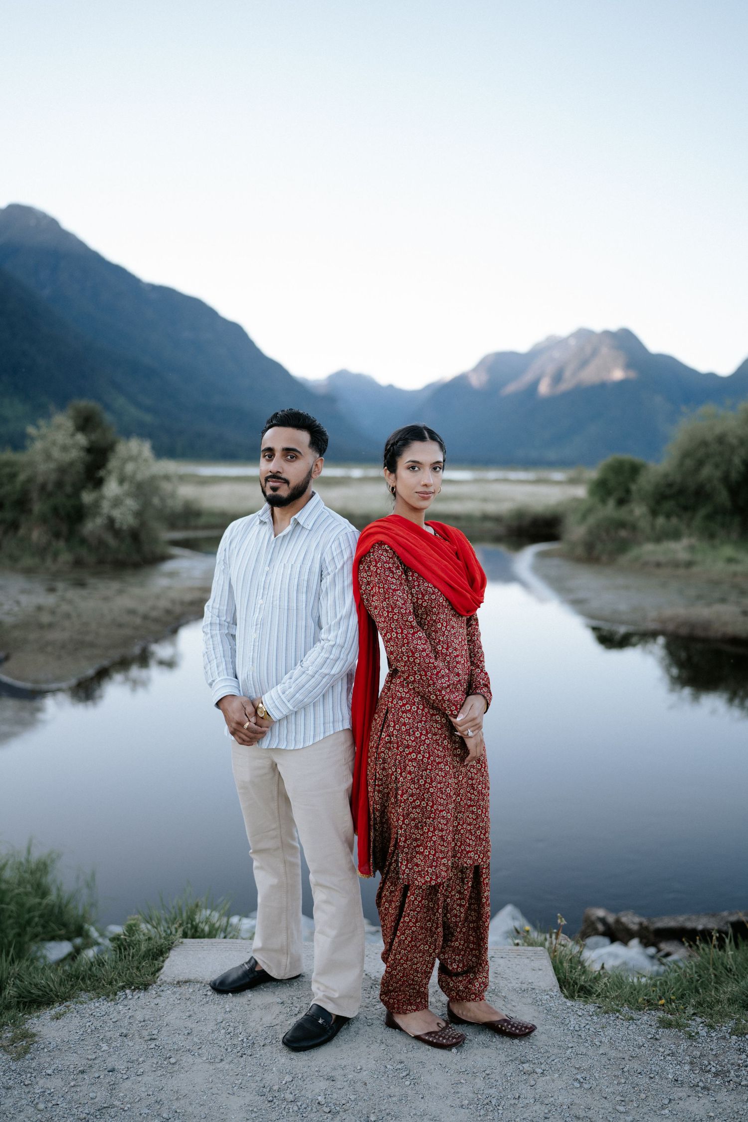 A couple poses for romantic photos by a serene mountain lake at dusk with scenic mountain peaks in the background.