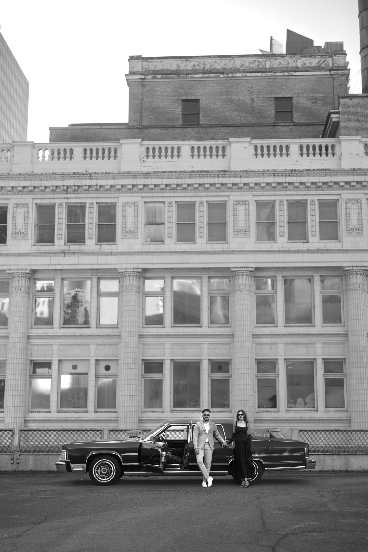 Black and white photograph of people standing near classic cars parked in front of historic brick buildings.