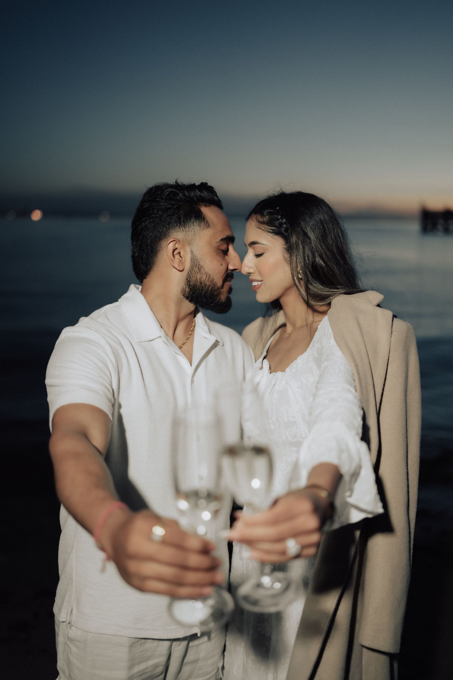 A romantic couple shares an intimate moment at sunset by the water while holding wine glasses.