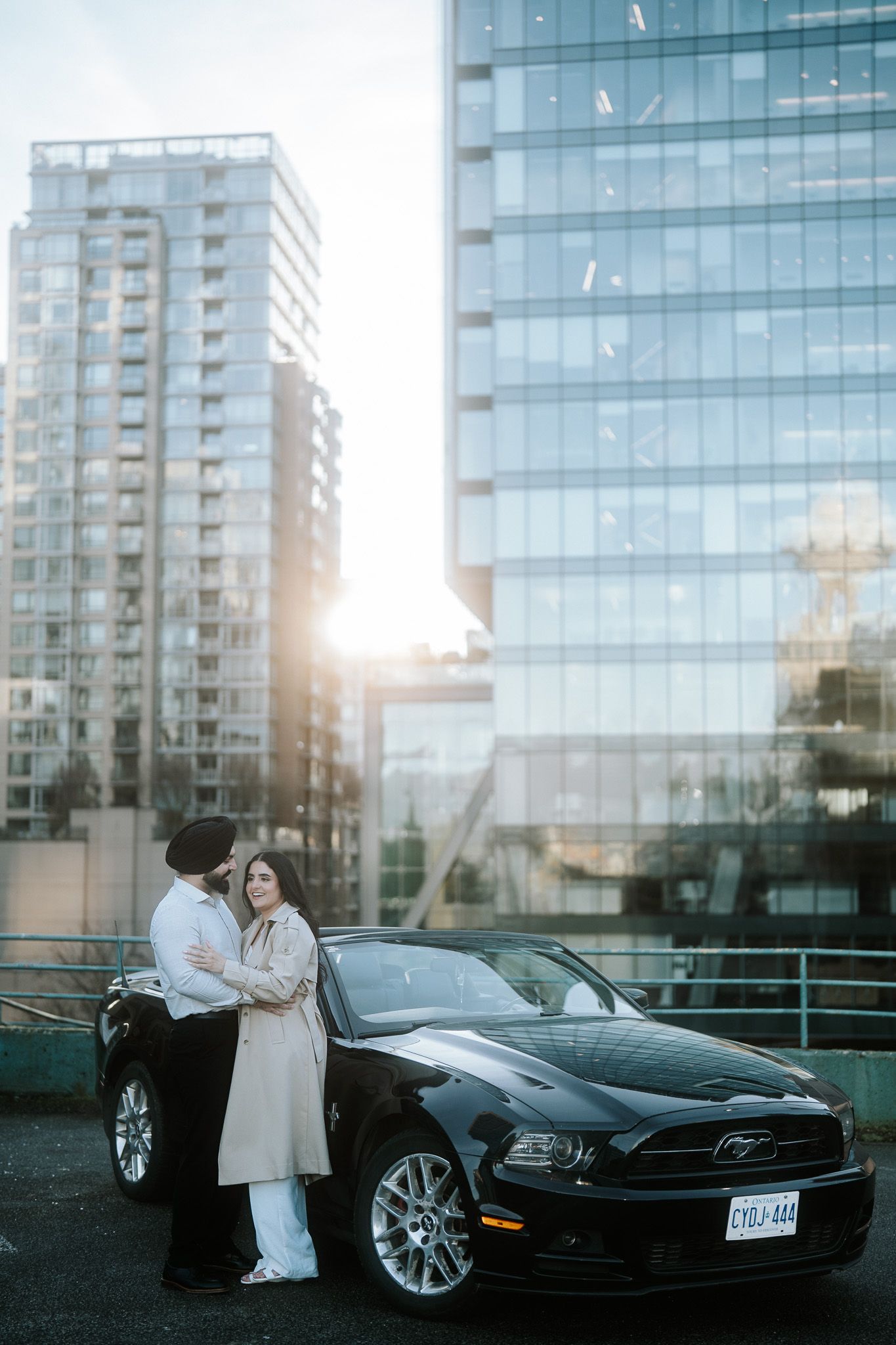 A couple stands beside a black Ford Mustang at sunset with modern glass buildings in the urban background.