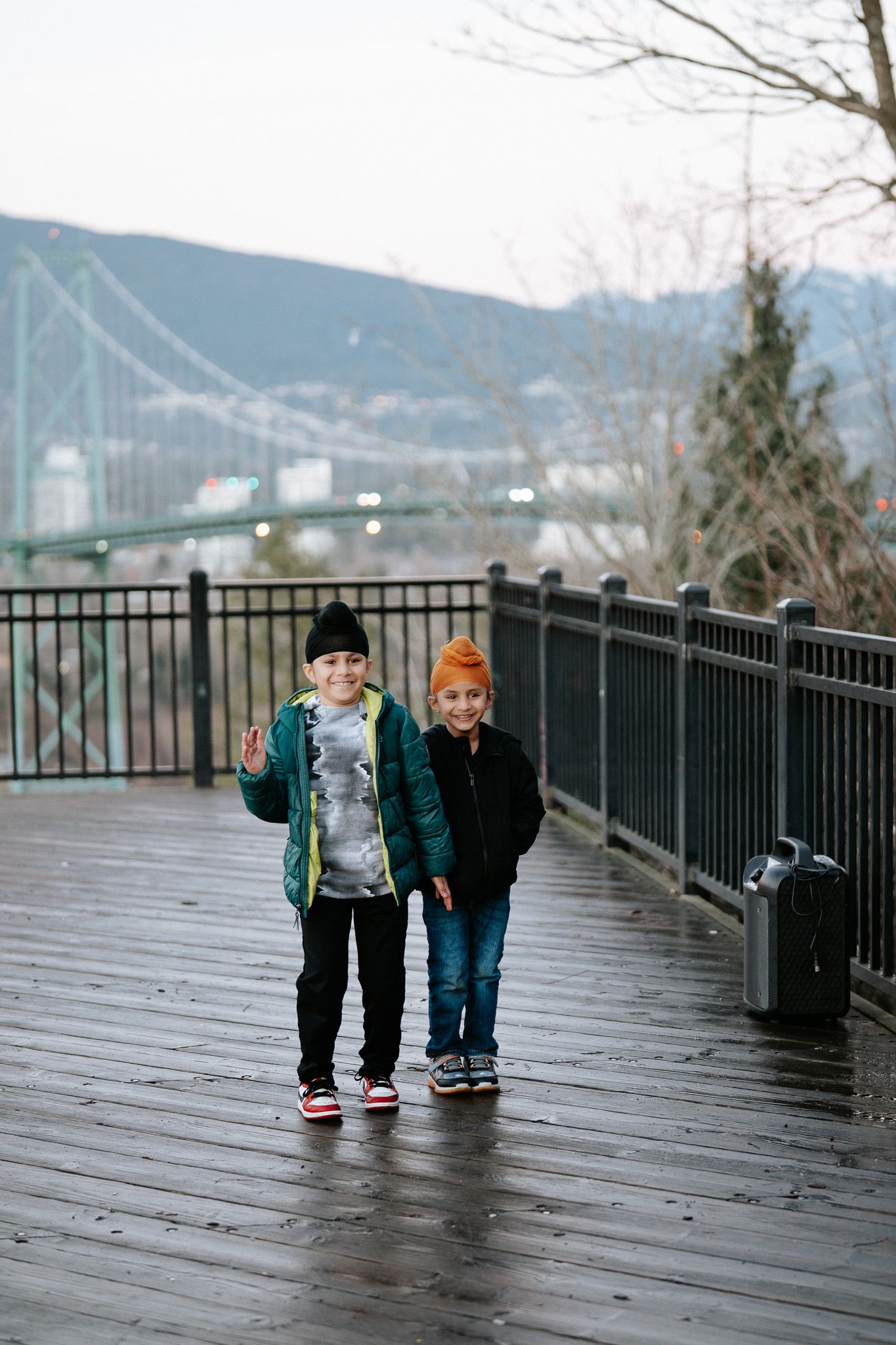 Two small children walk together on a wet wooden boardwalk with mountains visible in the background.