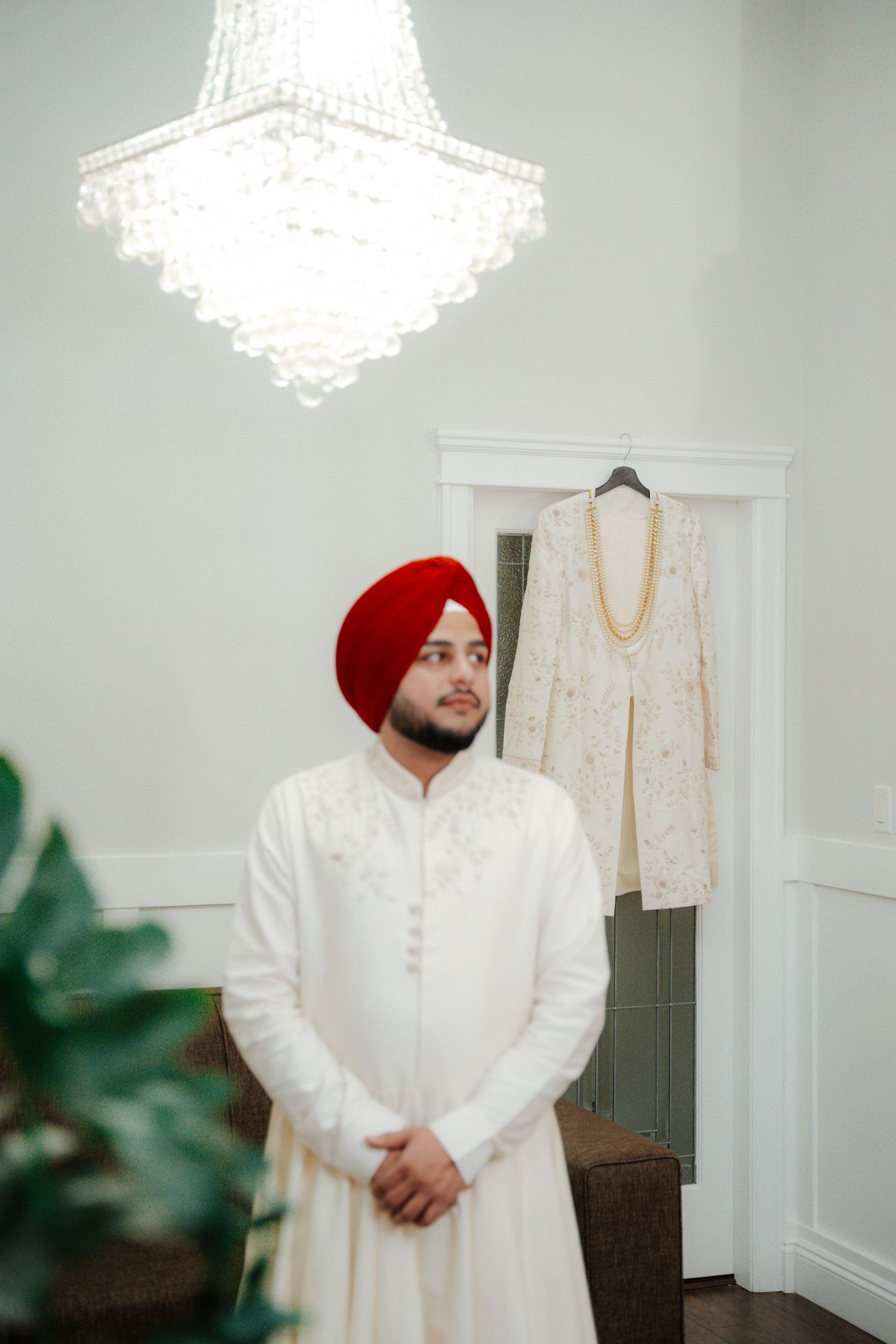 Traditional Sikh groom wearing a white sherwani and red turban standing in an elegant room with chandeliers.