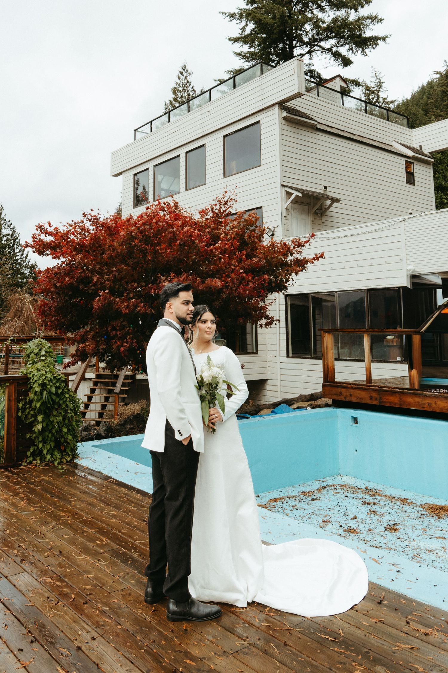 A couple in wedding attire stands on a wooden deck next to an empty pool and white house with vibrant red autumn foliage.