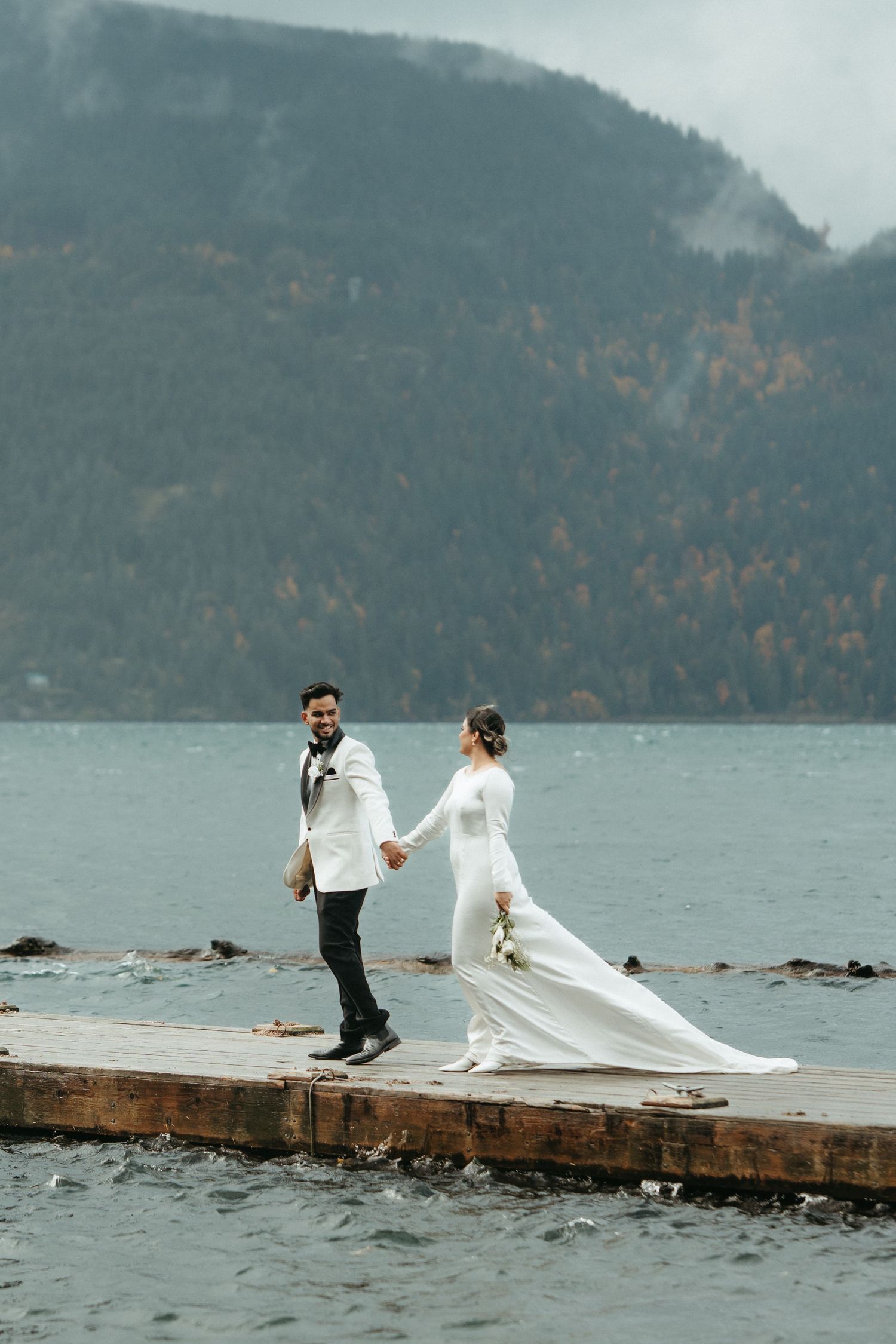 A couple in wedding attire poses dramatically on a wooden dock with misty mountains and lake in the background.