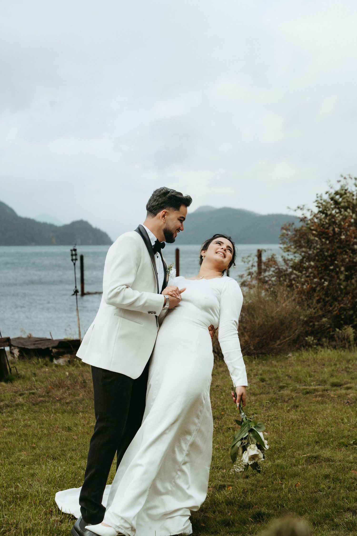 A couple in wedding attire share a romantic moment on a hilltop overlooking a misty bay and mountains.