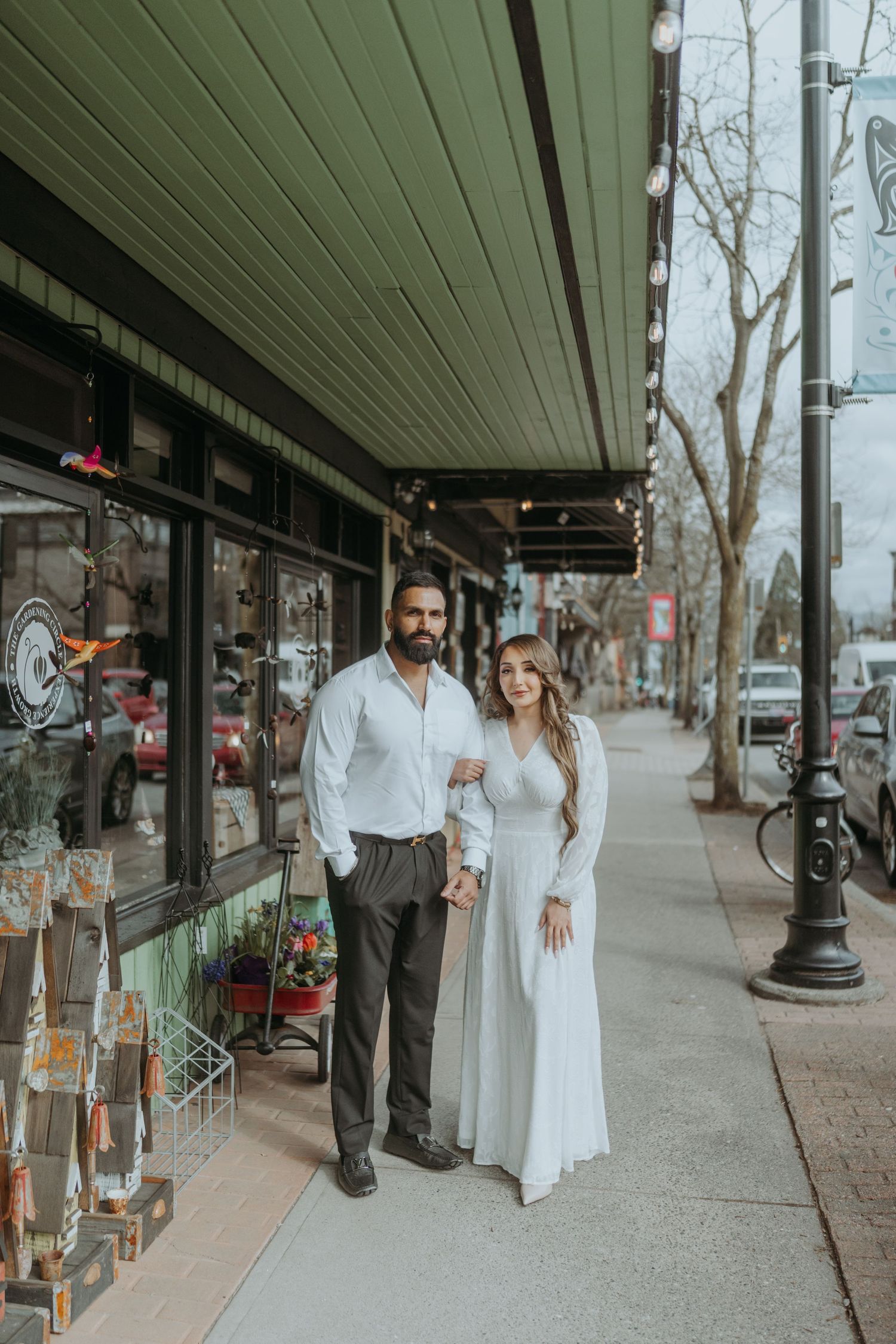 A couple stands together on a sidewalk in front of a shop with green awning and string lights.