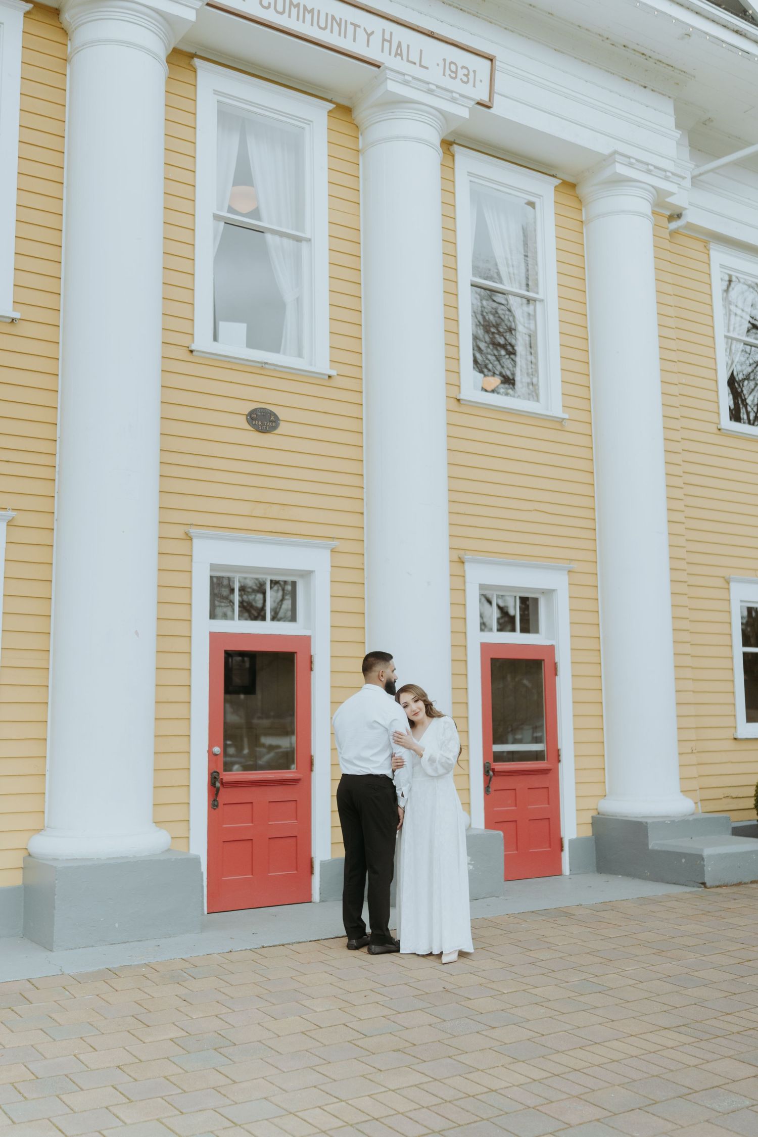 Wide shot of a couple posing against a yellow colonial building with red doors.