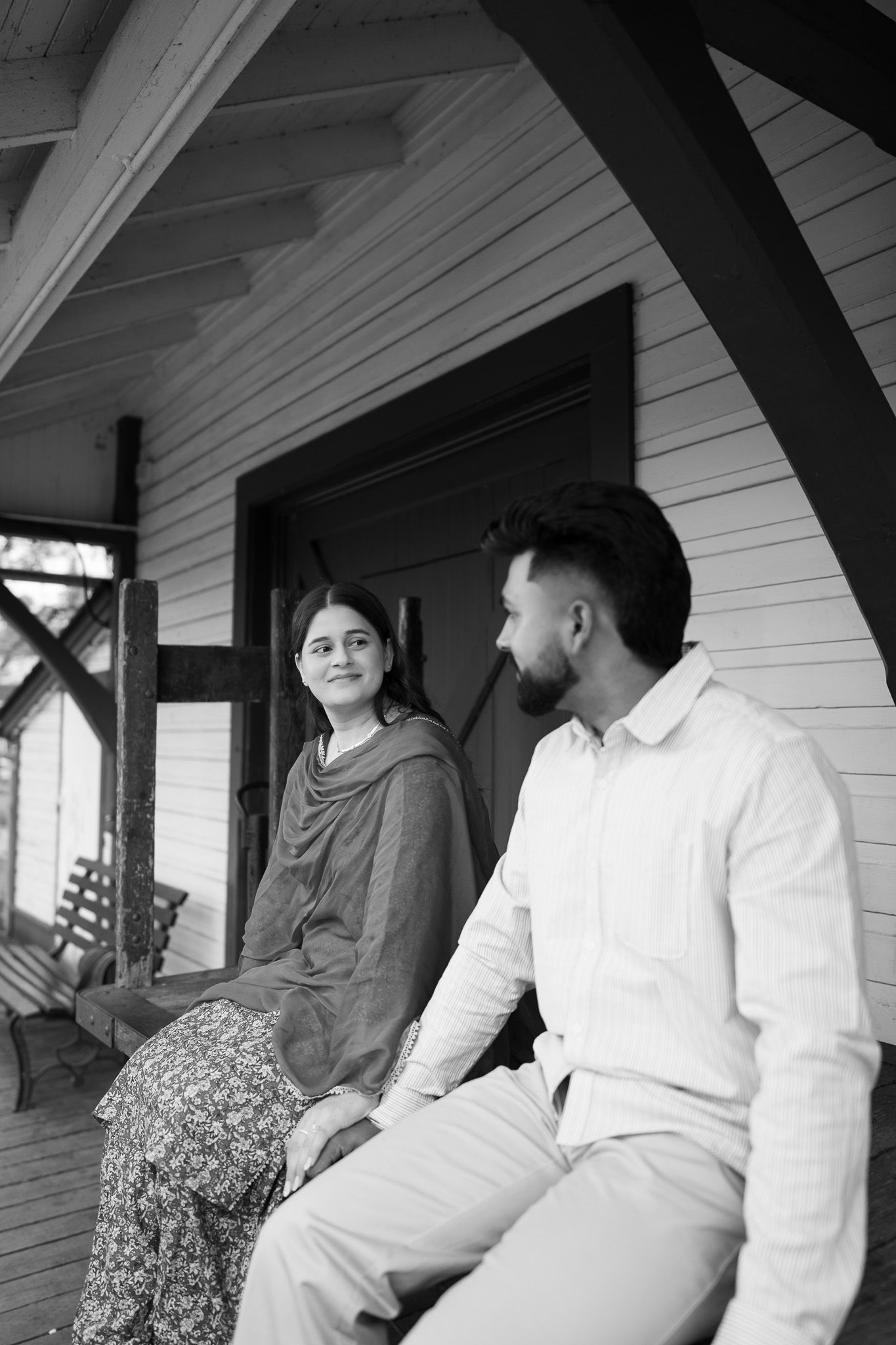 Black and white photo of a South Asian couple sitting on a porch, woman in dupatta smiling at bearded man.