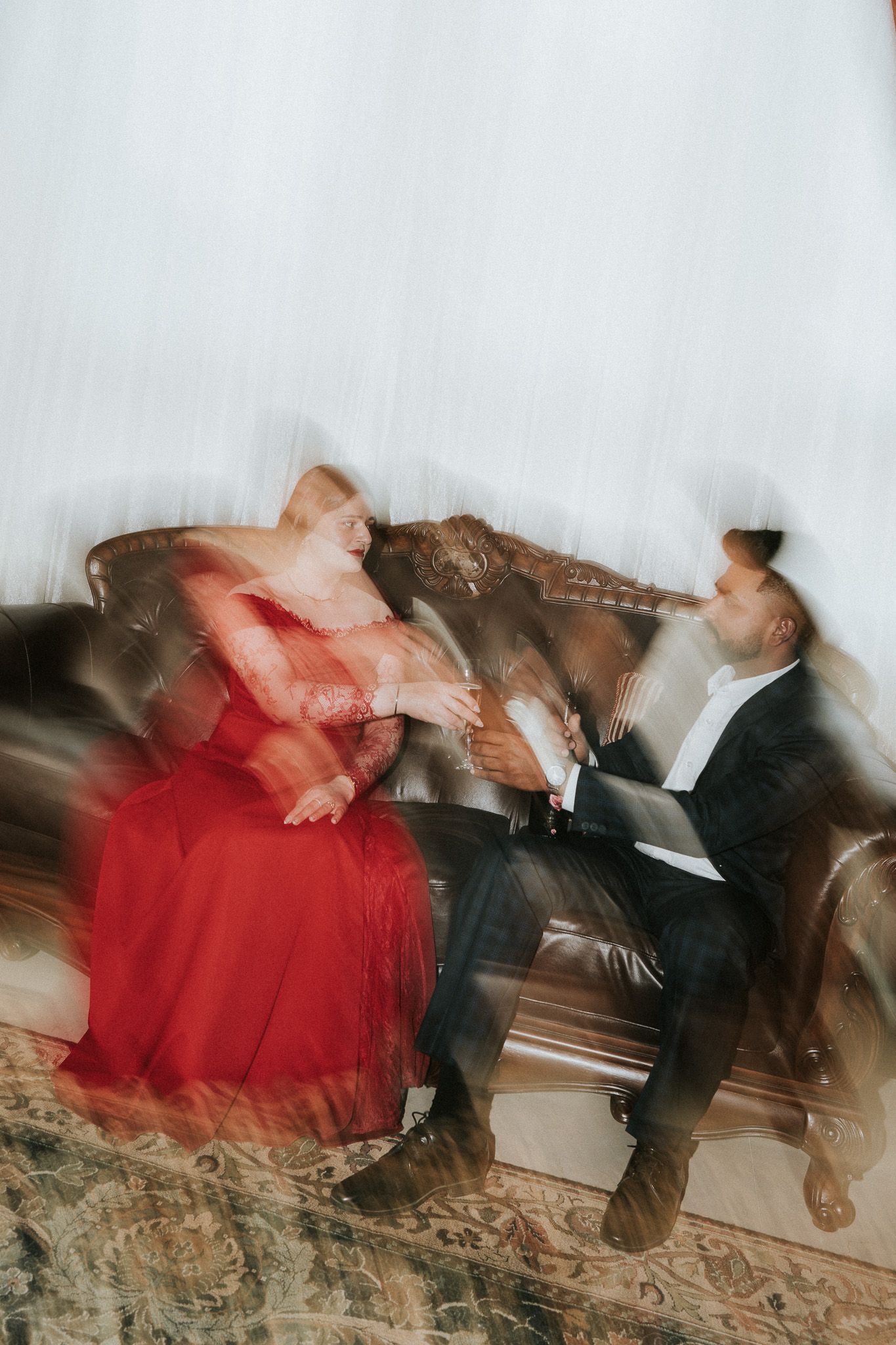 Motion blur artistic photo of couple in formal wear sharing champagne on a leather sofa.