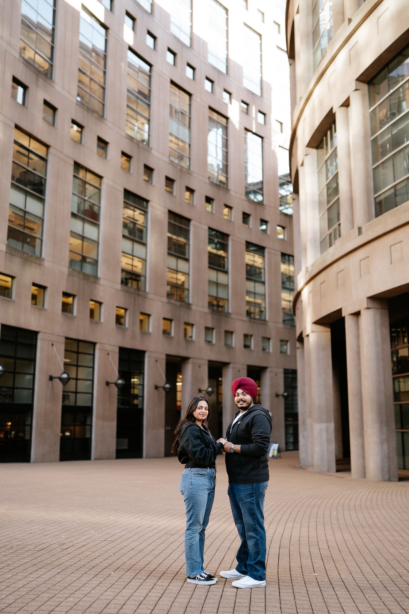 Two people stand together in a courtyard surrounded by modern curved architecture with tall windows.