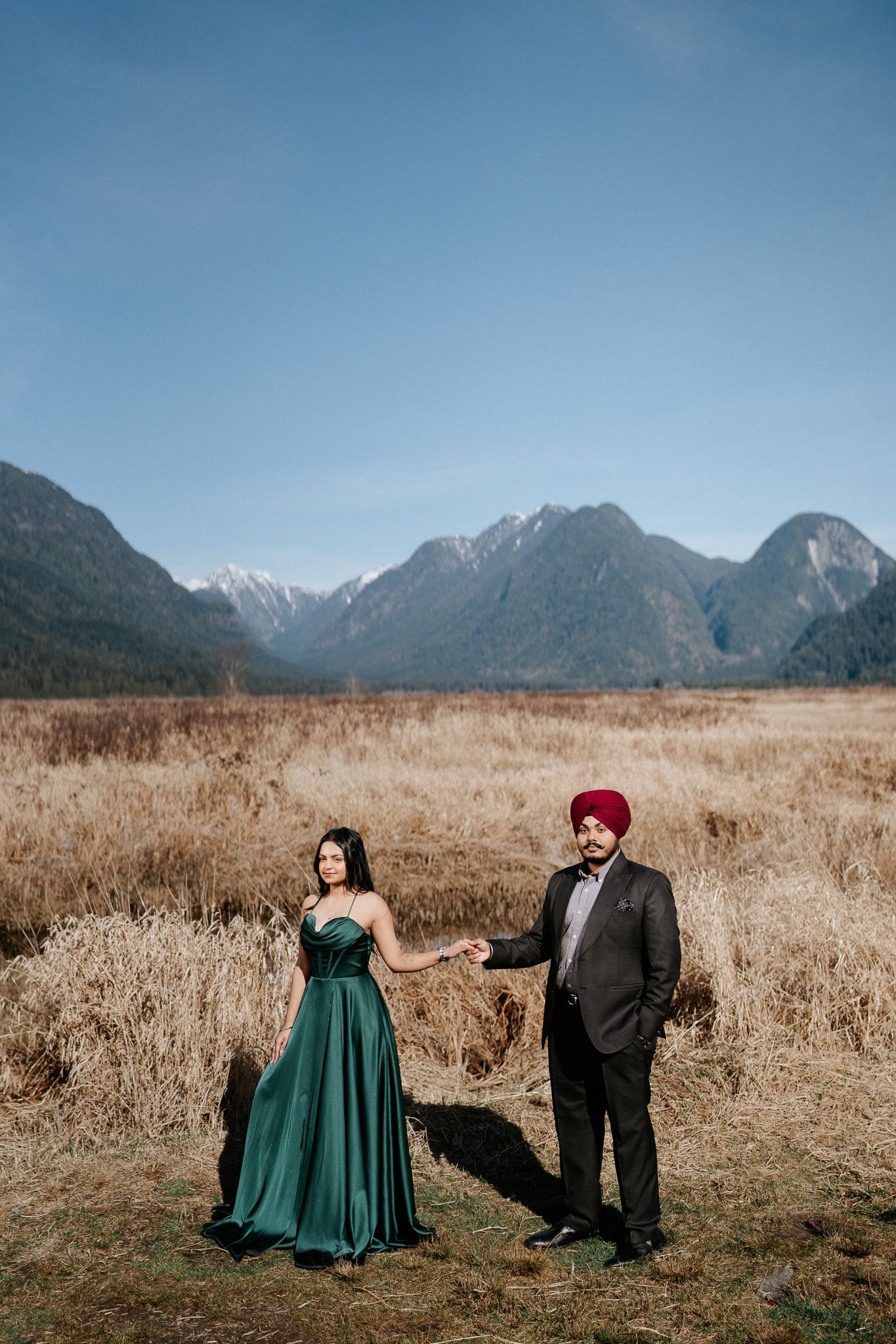Couple in formal attire poses in golden field with majestic mountain range backdrop during outdoor photo session.
