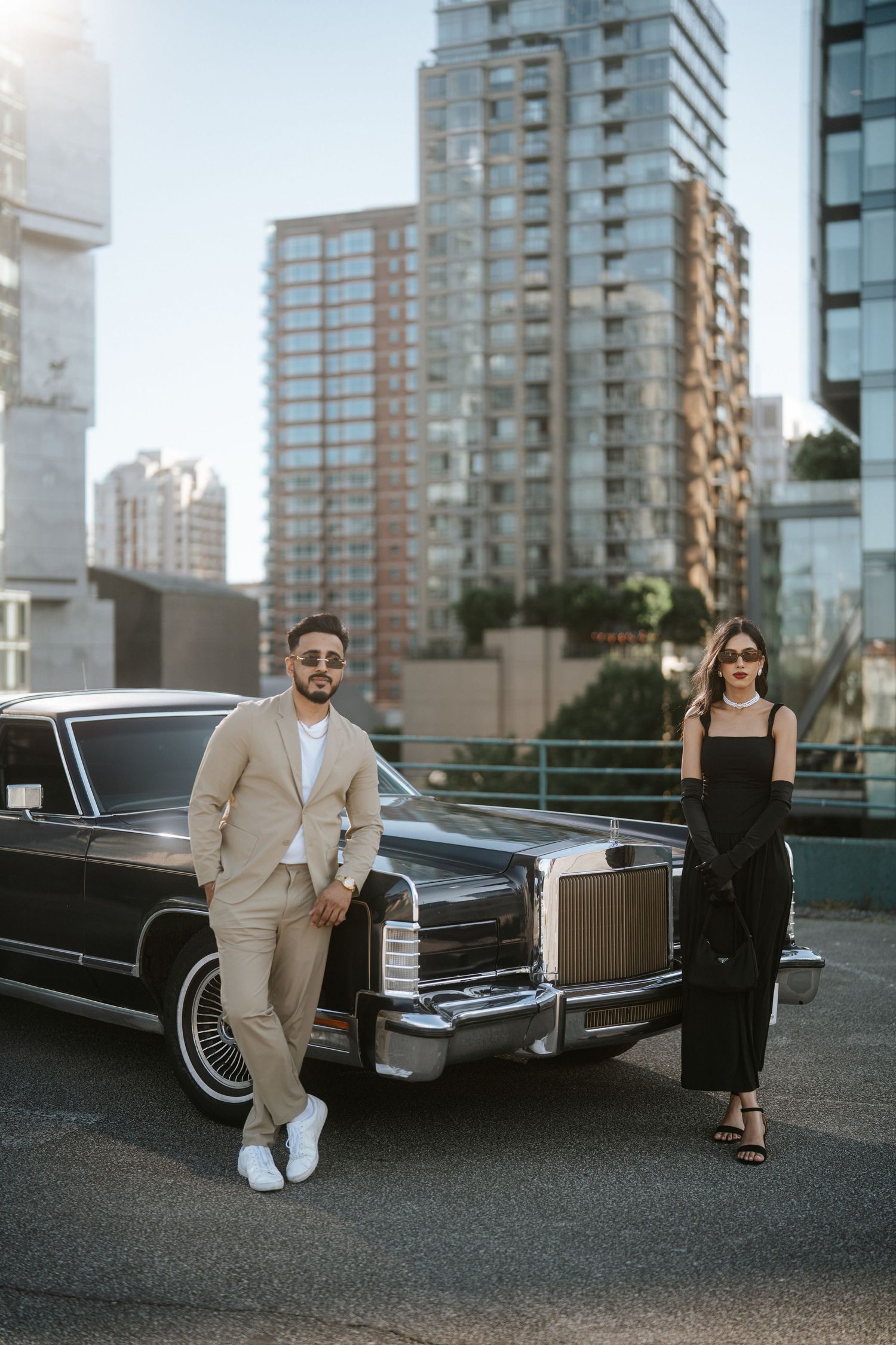 A couple poses elegantly next to a vintage black Lincoln Continental car with modern high-rise buildings in the background.