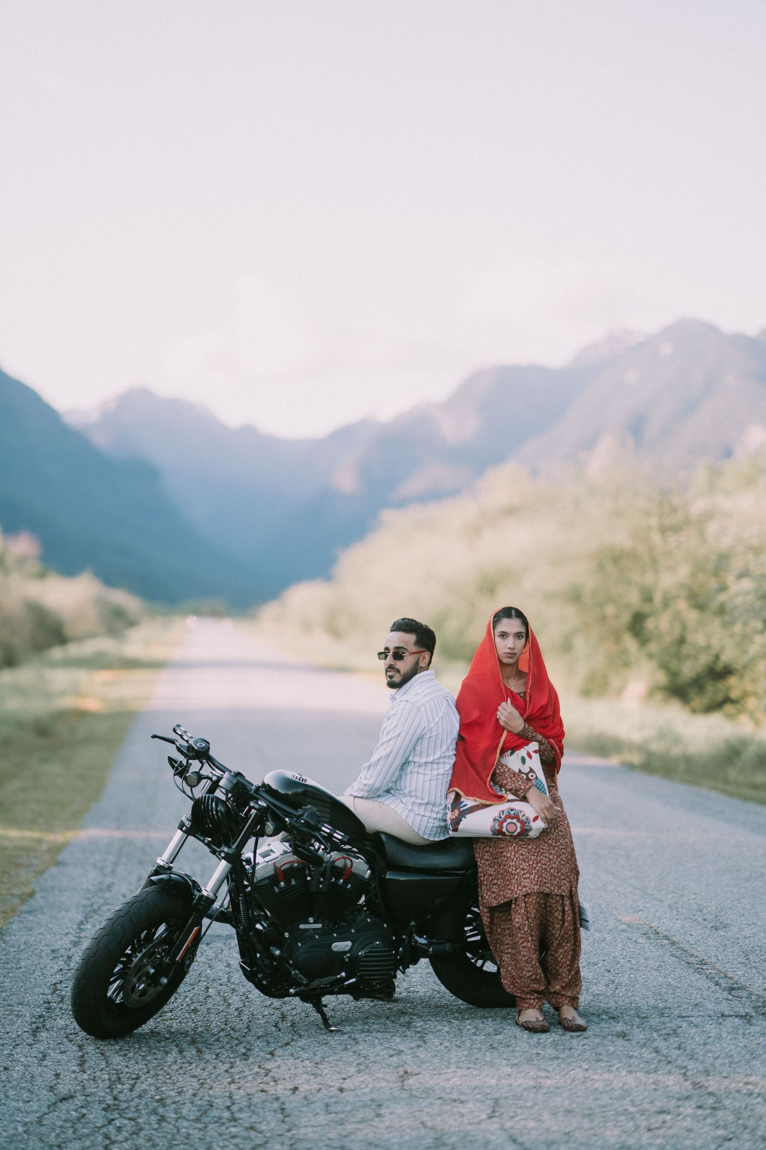 A motorcycle parked on a mountain road with two people posing beside it against a scenic mountain backdrop.