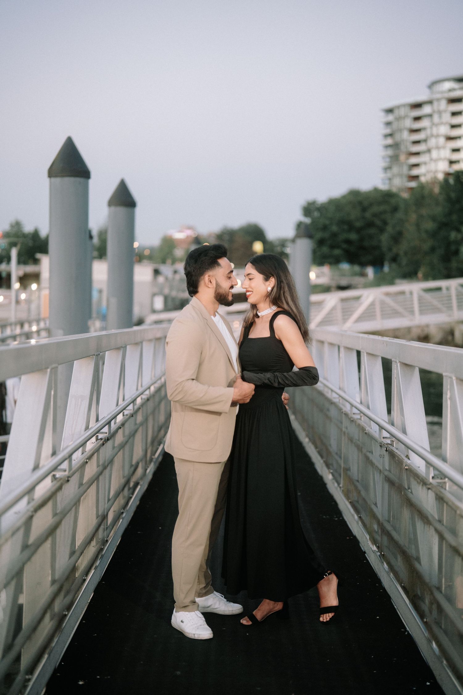 A romantic couple in contrasting black and beige outfits share intimate moments on a modern white bridge walkway.
