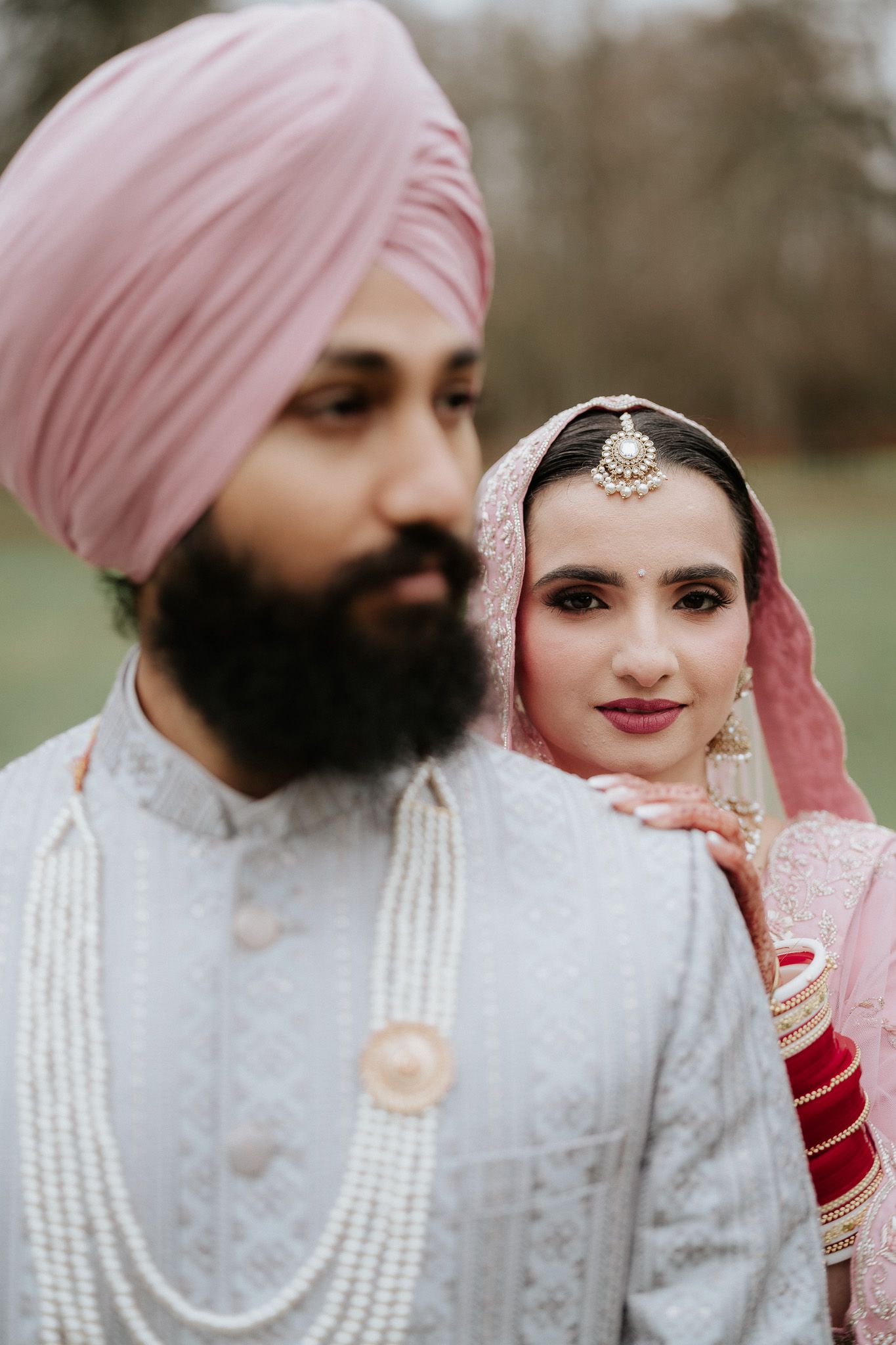 A couple in traditional Sikh wedding attire pose together outdoors, with the bride in a pink dupatta and groom in a pink turban.