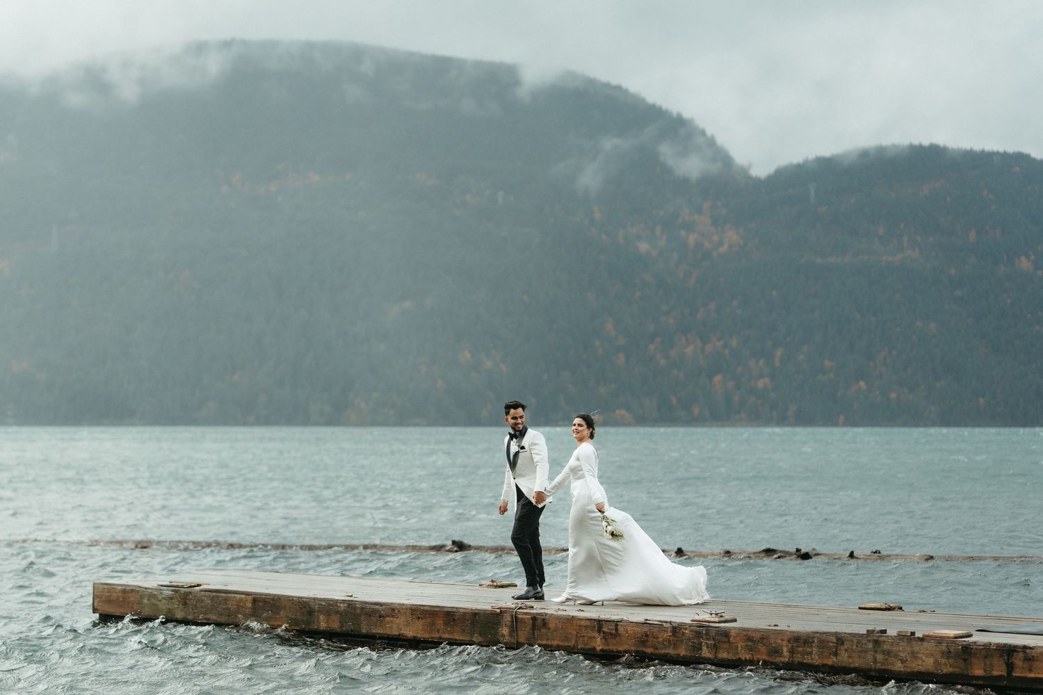 A couple in wedding attire stands on a misty pier overlooking a serene mountain lake on their special day.