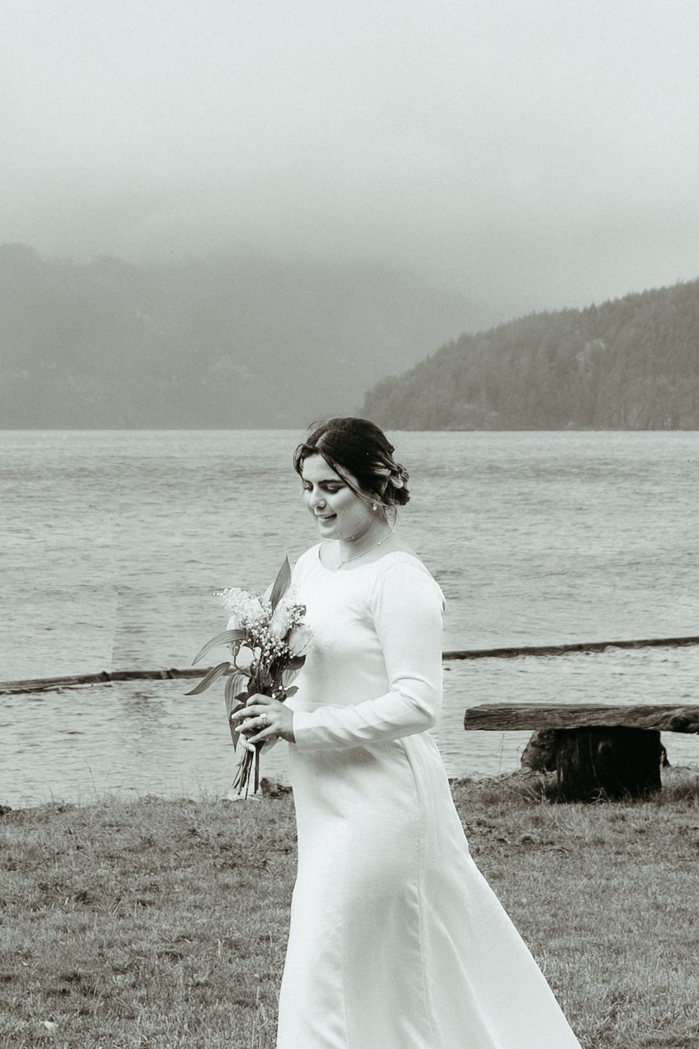 A person in a white wedding dress holds a bouquet while standing near a misty lake with mountains in the background.
