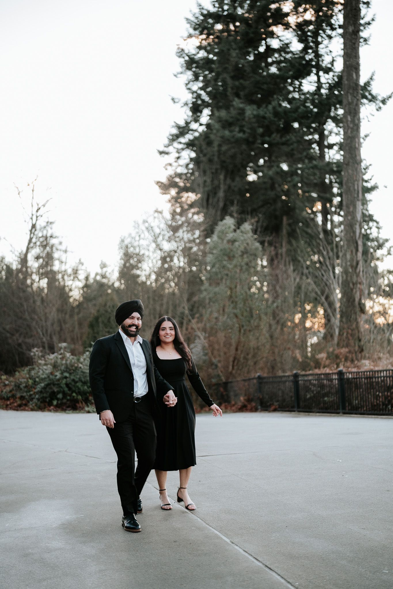 A couple in formal attire playfully poses on a concrete surface surrounded by evergreen trees at dusk.