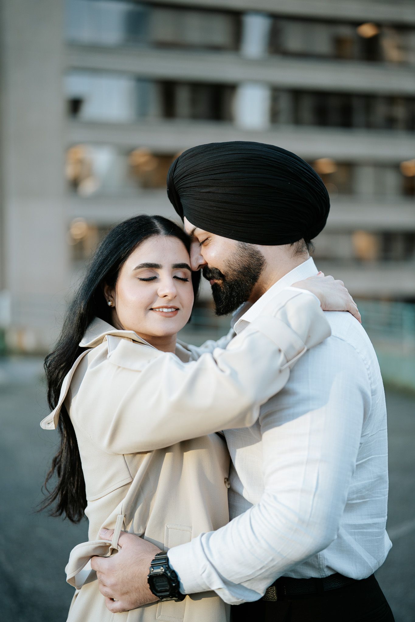 A loving couple embraces outdoors, with one wearing a turban and white shirt while the other wears a beige coat.