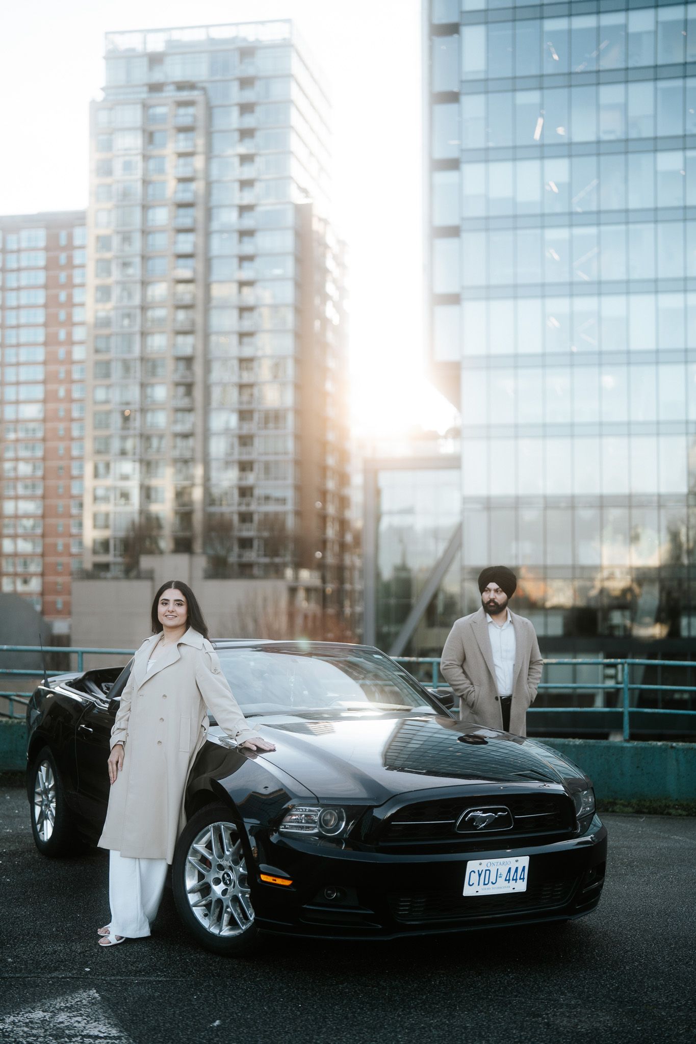 Stylish couple poses with black Ford Mustang in front of modern high-rise buildings during sunset.
