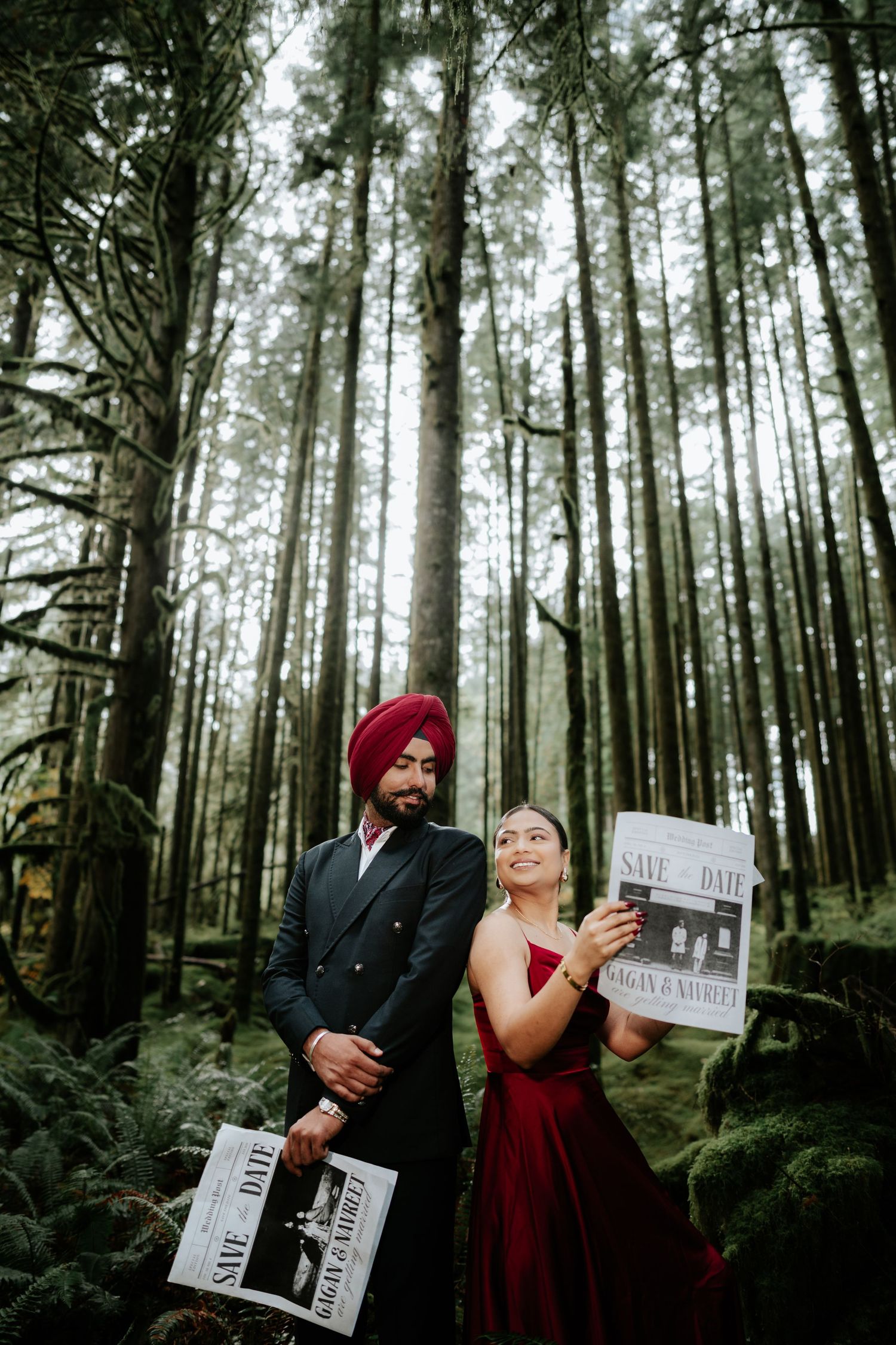 A couple in formal wear shares save-the-date newspapers in a lush forest setting.