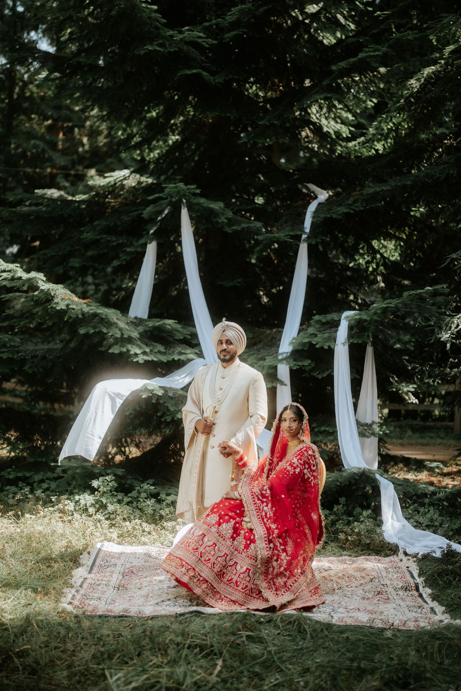 A wedding couple sits on a rug outdoors with white fabric draping in the background.