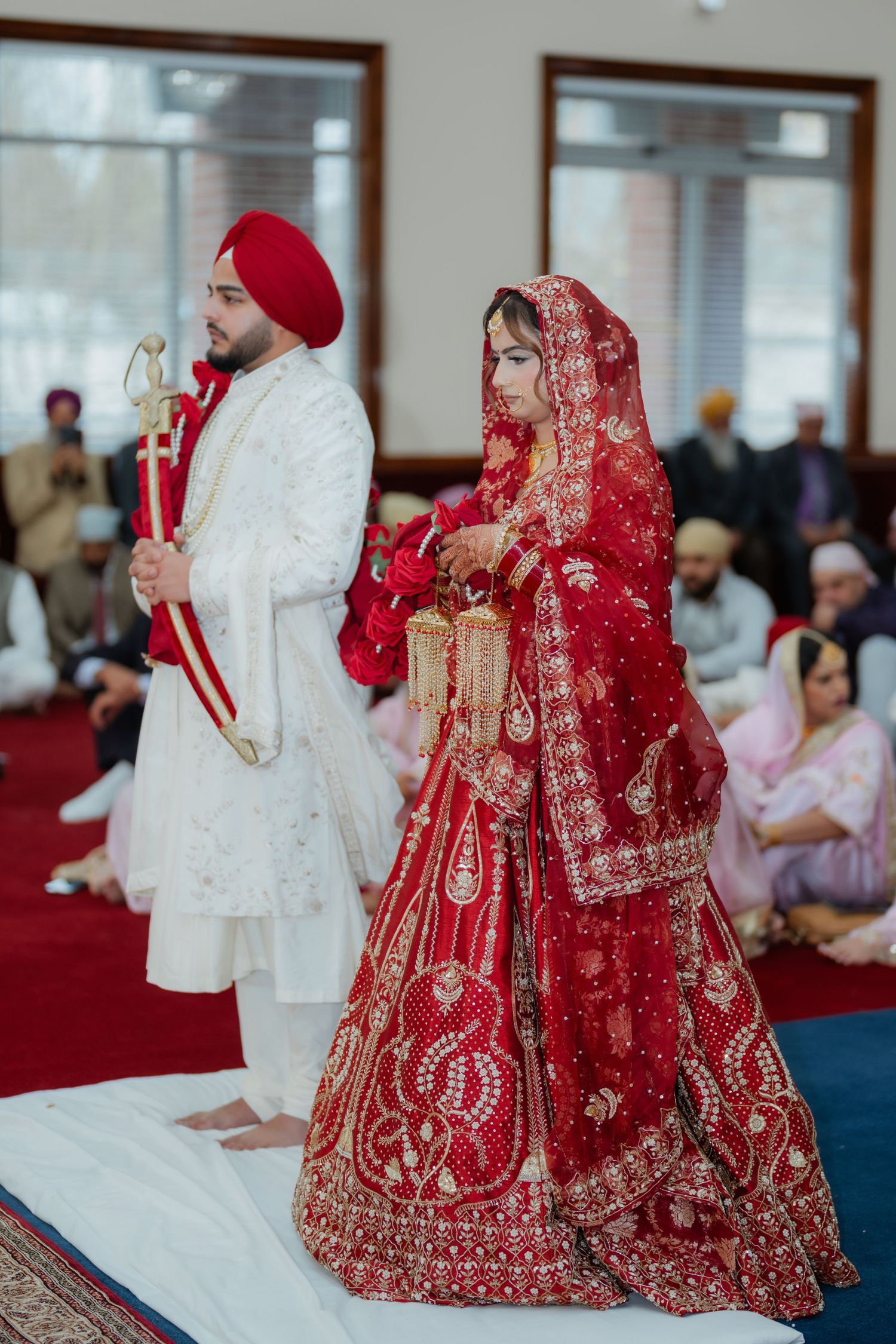 Traditional Sikh wedding ceremony with attendees in formal attire seated on blue and red carpeted floor.