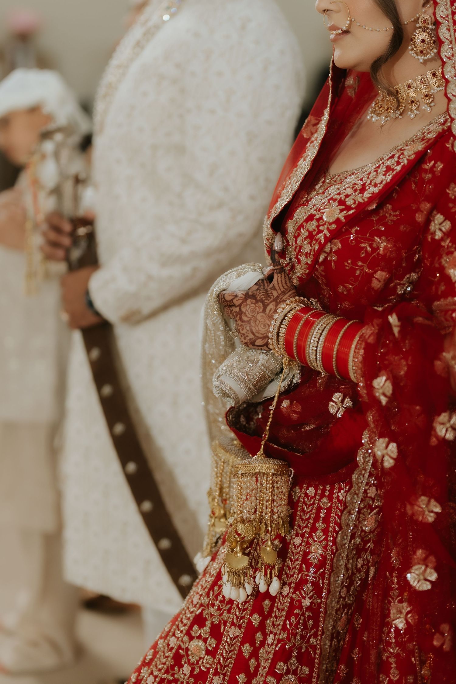 Indian bride in red embroidered lehenga with gold jewelry and bangles holds ornate purse at traditional wedding ceremony.