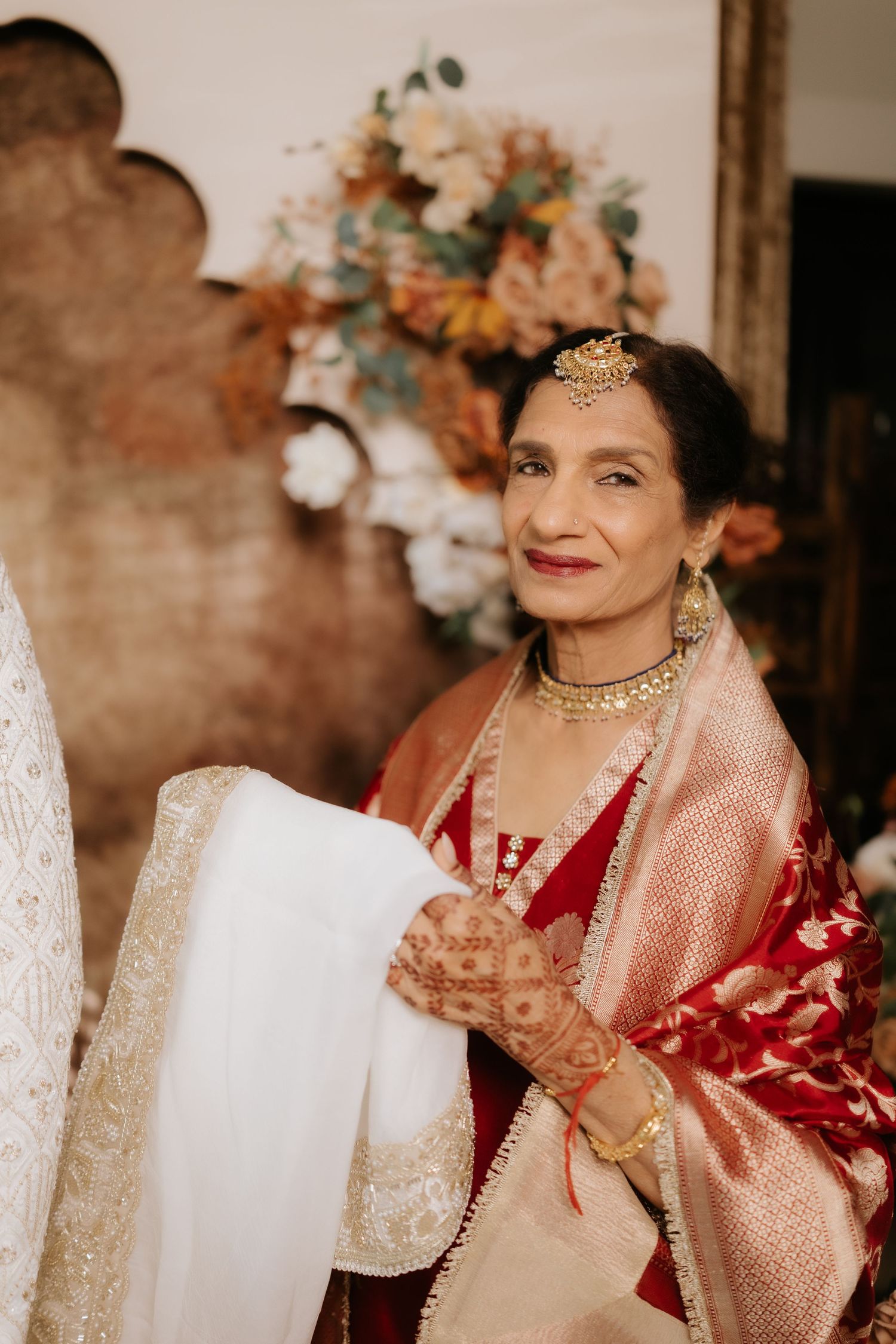 Elegant Indian woman in red and gold saree with traditional jewelry smiling at a wedding ceremony.