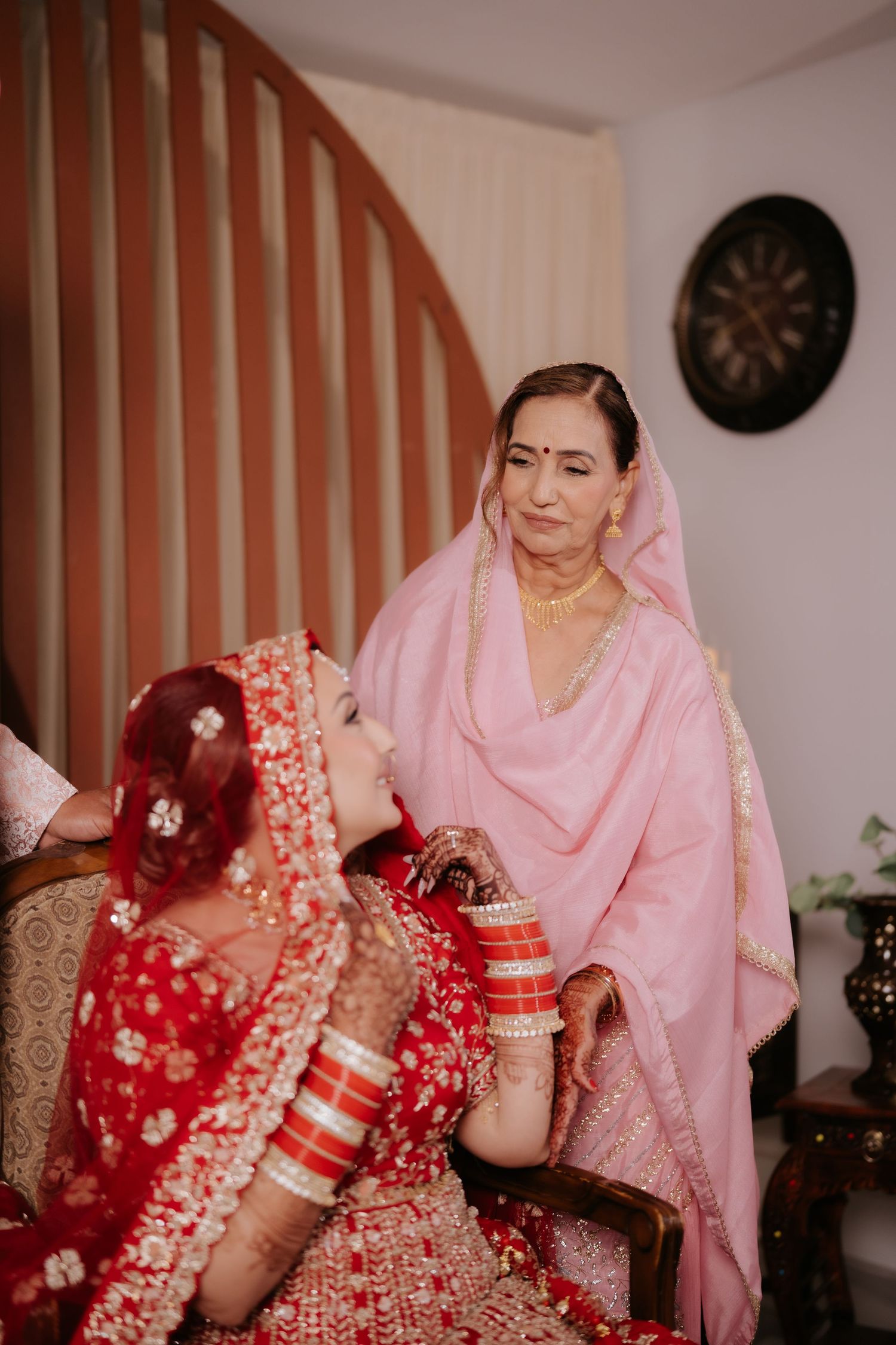 Indian bride in red lehenga looks up at elderly woman in pink salwar suit during pre-wedding ceremony indoors.