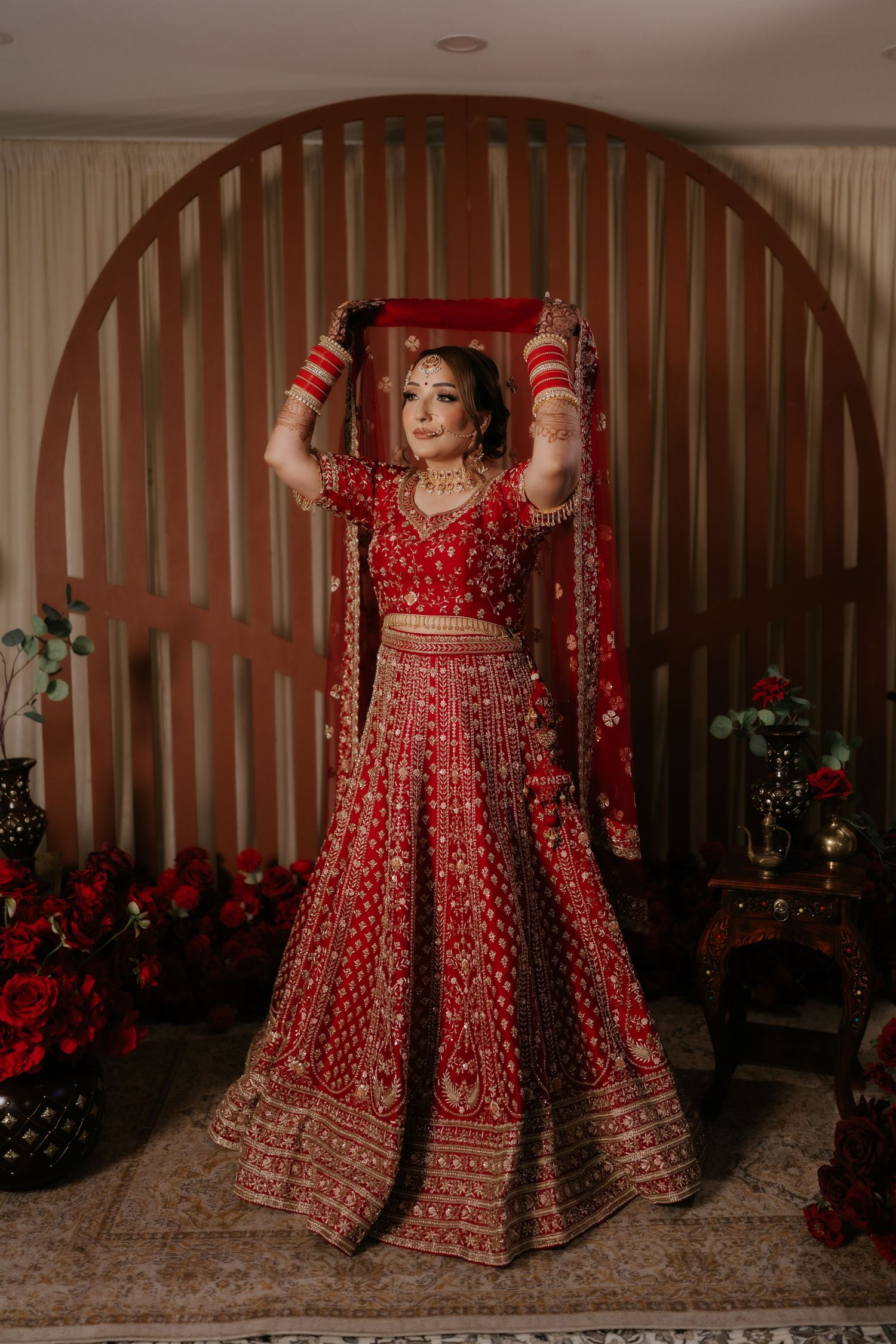 Bride in red embroidered lehenga holding dupatta overhead, posing before arched wooden backdrop with red floral decor.