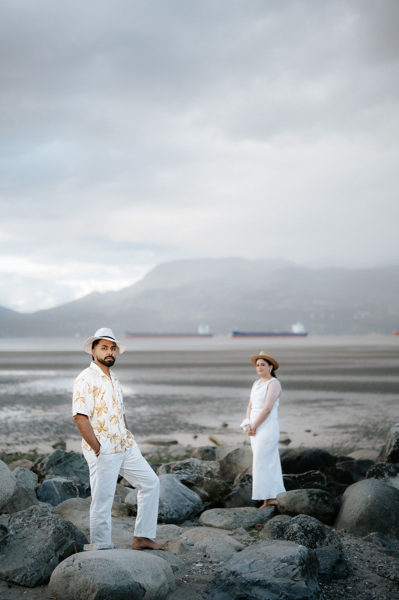 Couple in white outfits and hats standing on rocky beach with mountains and cargo ship in background.