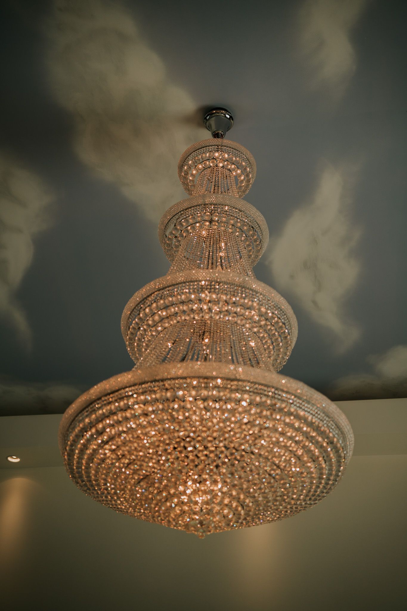 Ornate multi-tiered crystal chandelier hanging from a painted cloud ceiling in an elegant room.