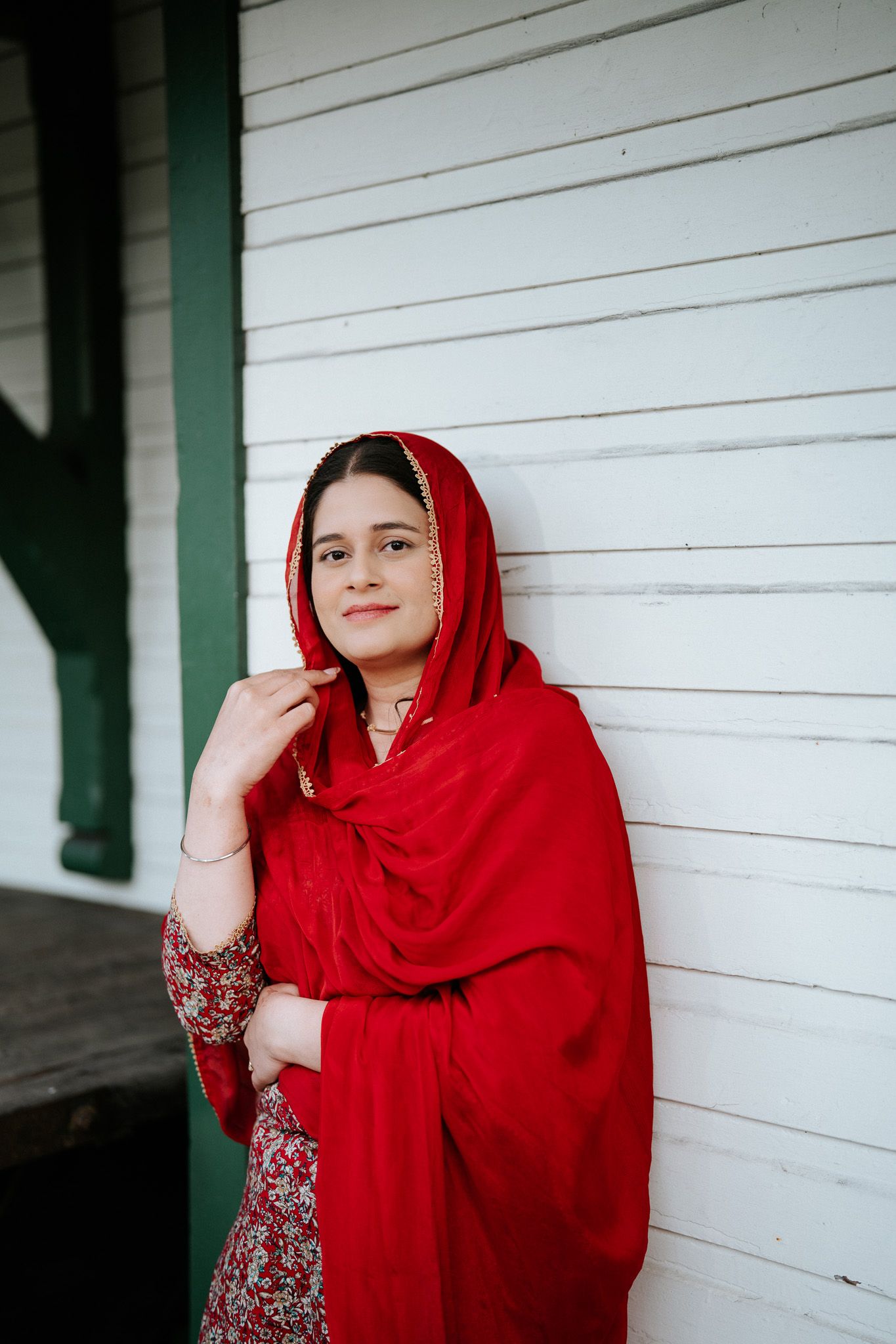 Portrait of a young South Asian woman in a vibrant red dupatta and floral dress leaning against a white wall.