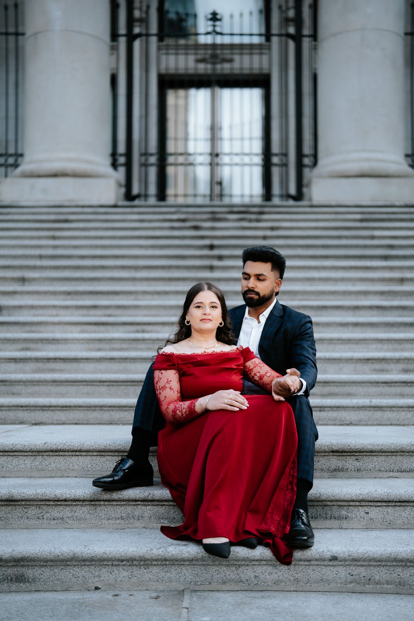 Elegant couple sitting on grand stone steps, woman in red lace gown, man in dark suit embracing her romantically.