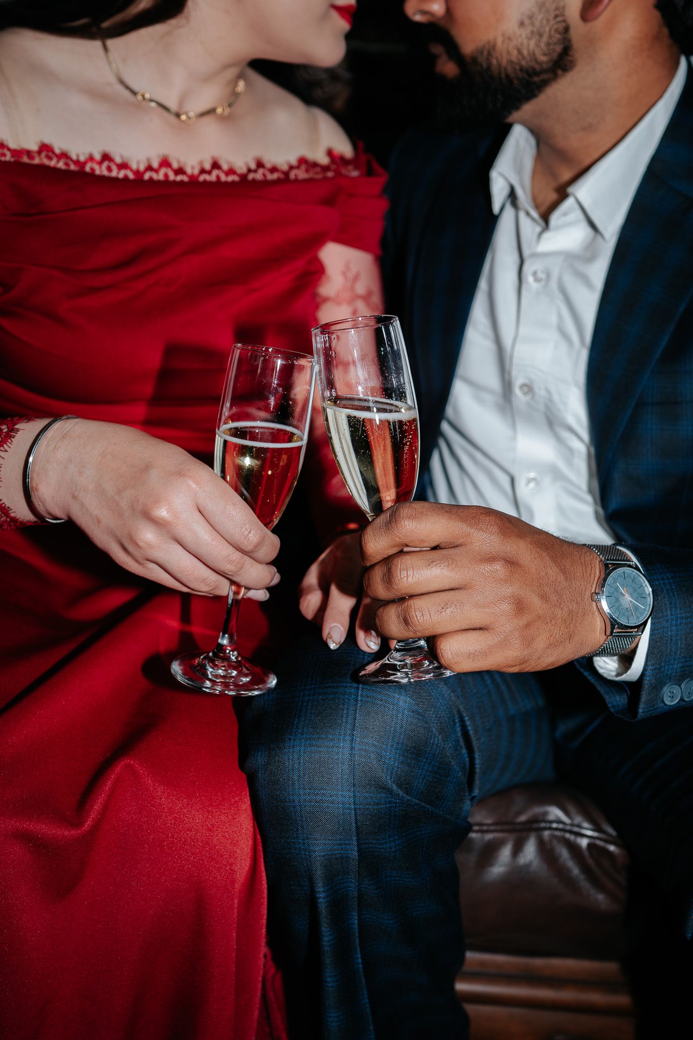 Couple clinking champagne glasses, woman in red off-shoulder dress and man in navy suit, romantic celebration.