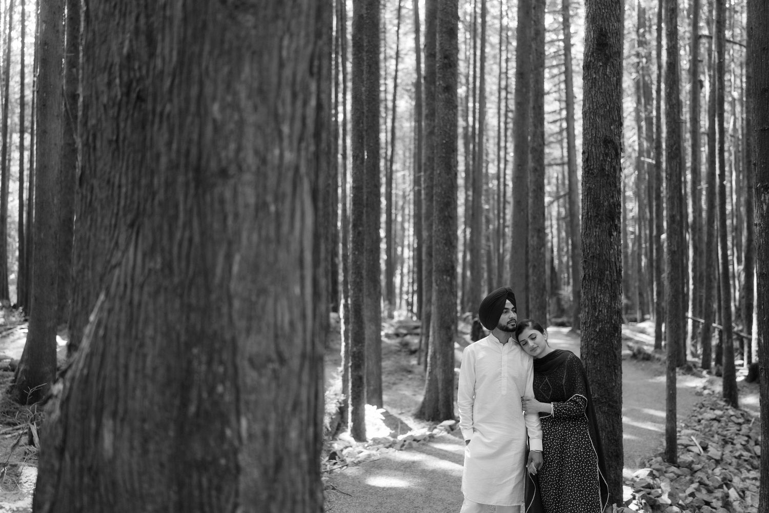 A couple in traditional Sikh attire walks through a tall forest in a romantic black and white photo.