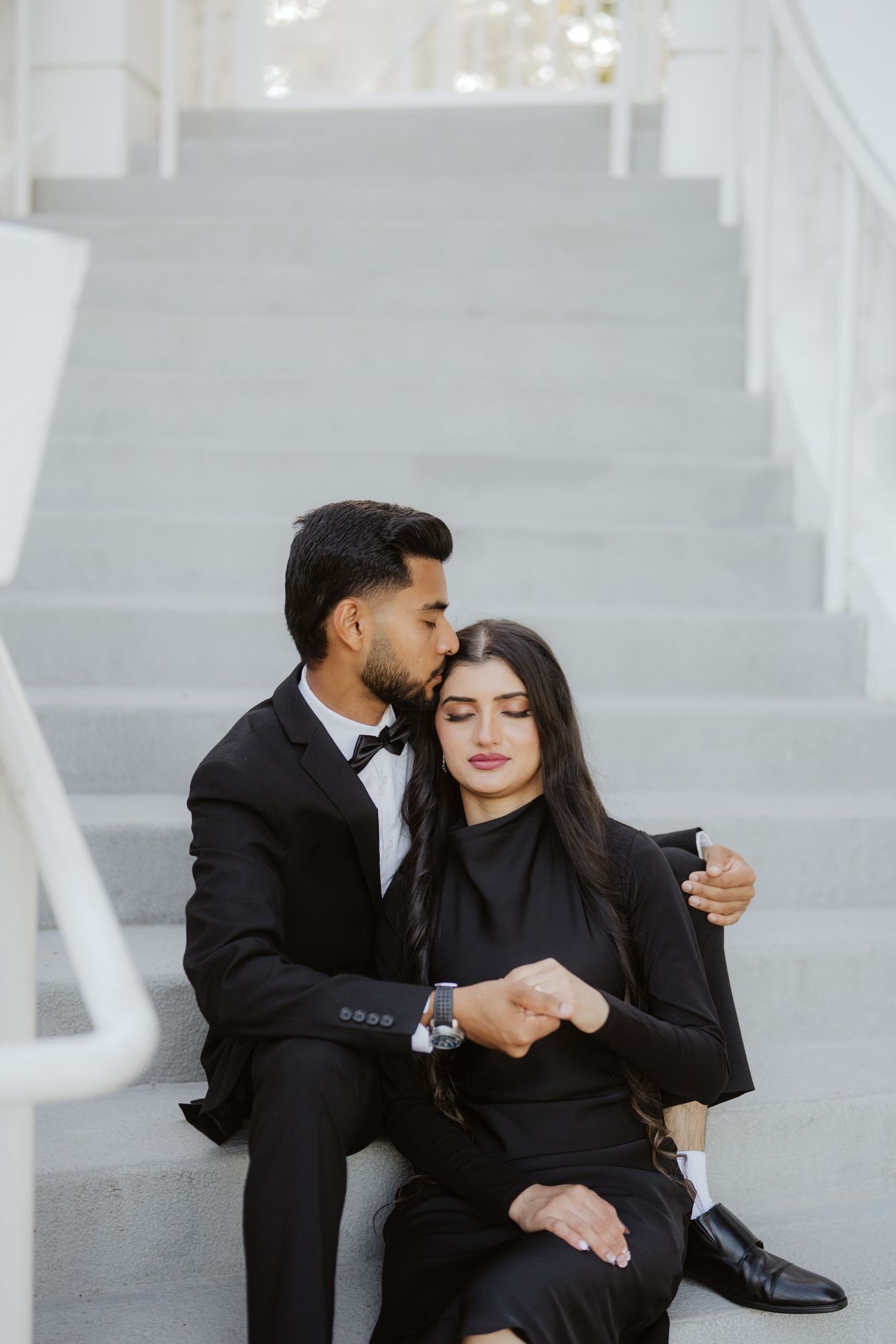 Man in black tuxedo kissing woman's head while sitting together on white marble steps outdoors.