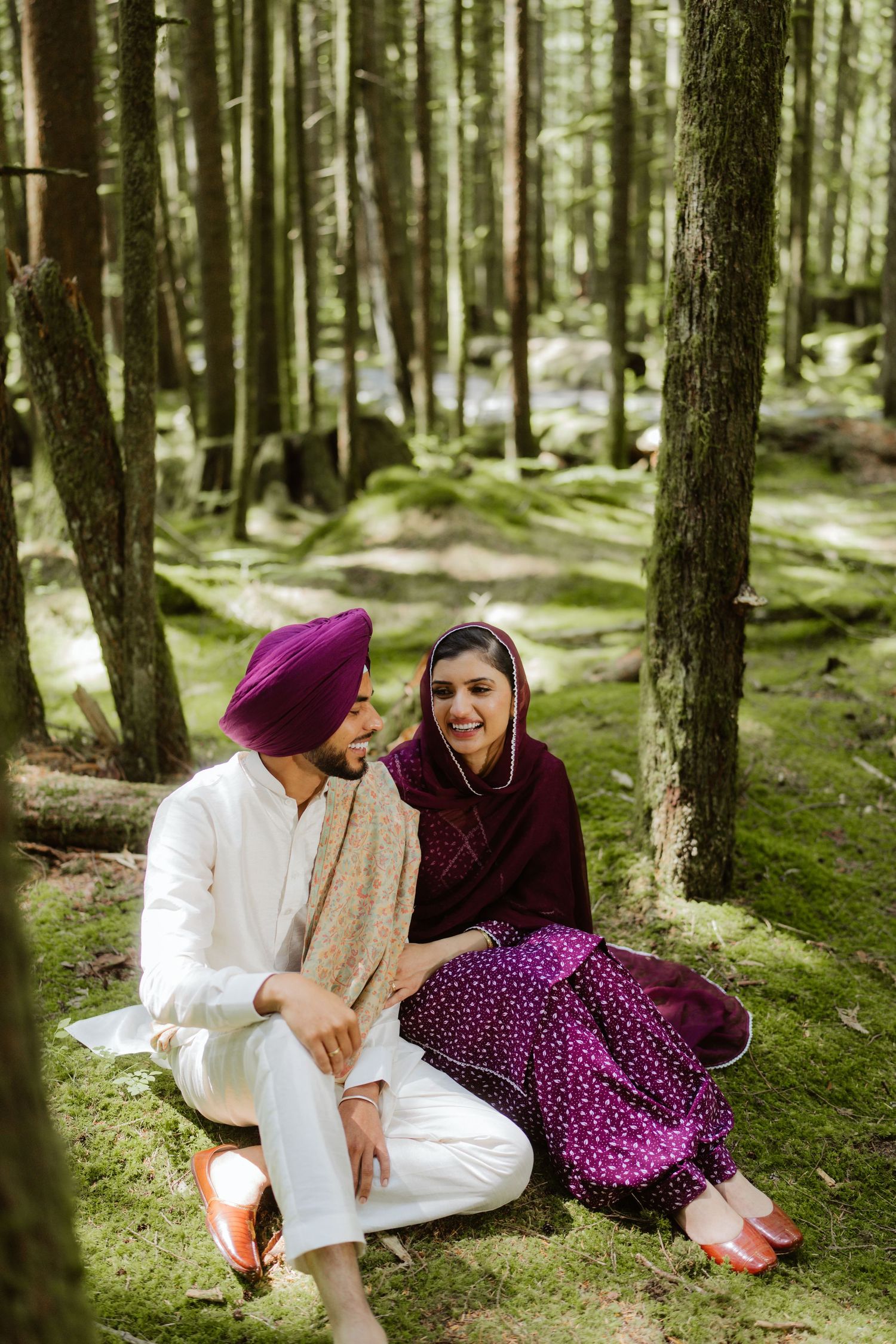 Sikh couple in traditional attire sit together on mossy forest floor, smiling and sharing a joyful moment.