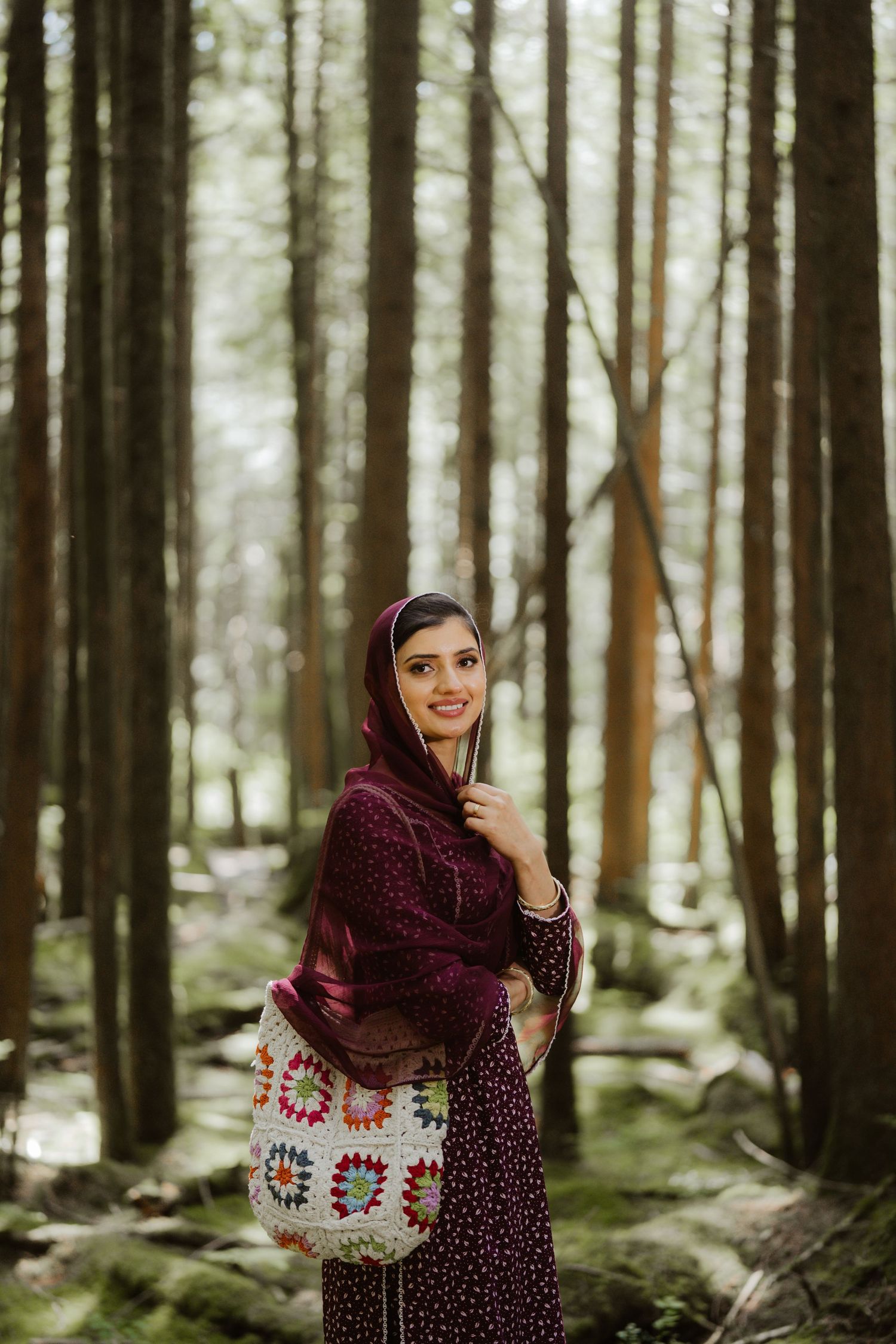 A smiling woman in a burgundy hijab and floral dress holds a colorful crochet bag while standing in a tall forest.